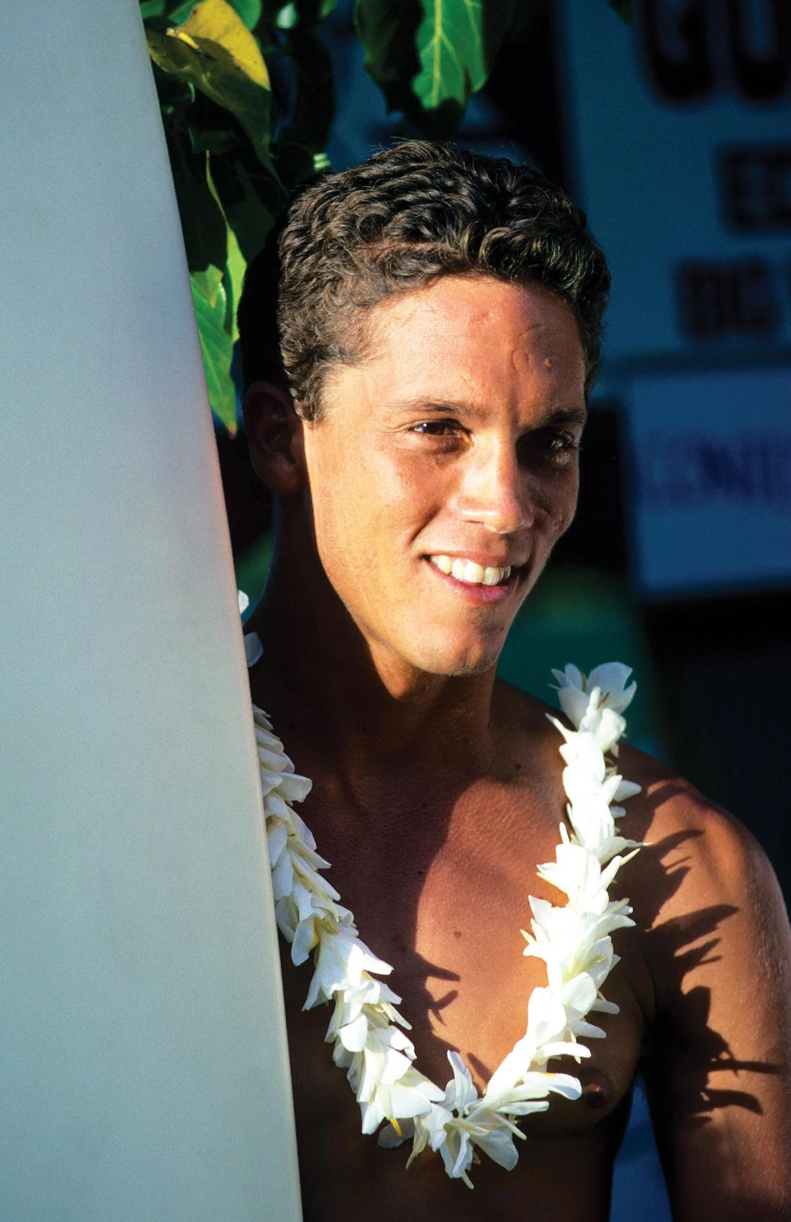 A shirtless man with curly dark hair smiling, wearing a necklace and flower lei, standing outdoors in bright sunlight, with green foliage in the background.