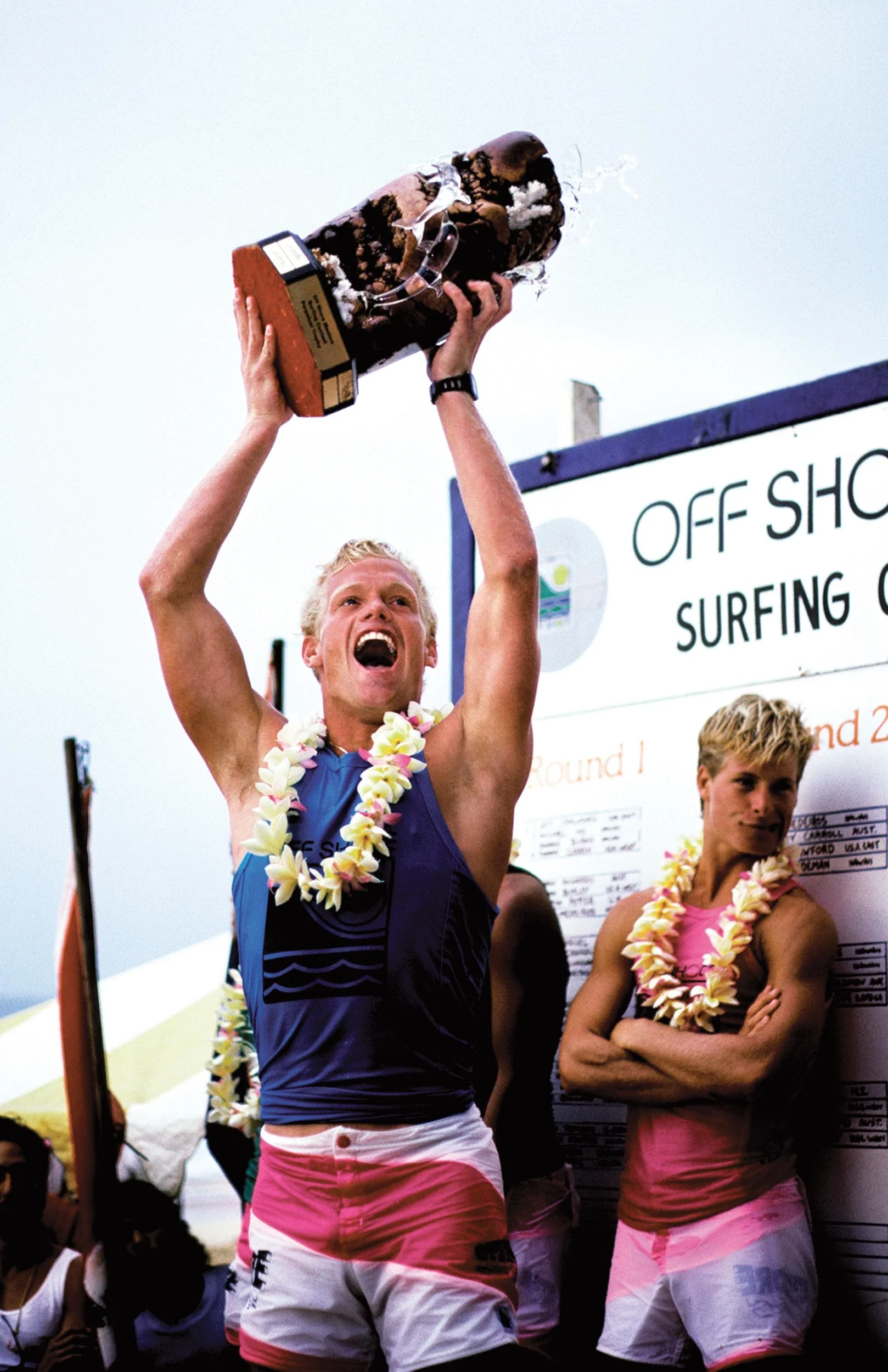 A man celebrating winning a surfing competition, holding a trophy above his head, wearing a floral lei and colorful shorts, with other competitors and a scoreboard in the background.