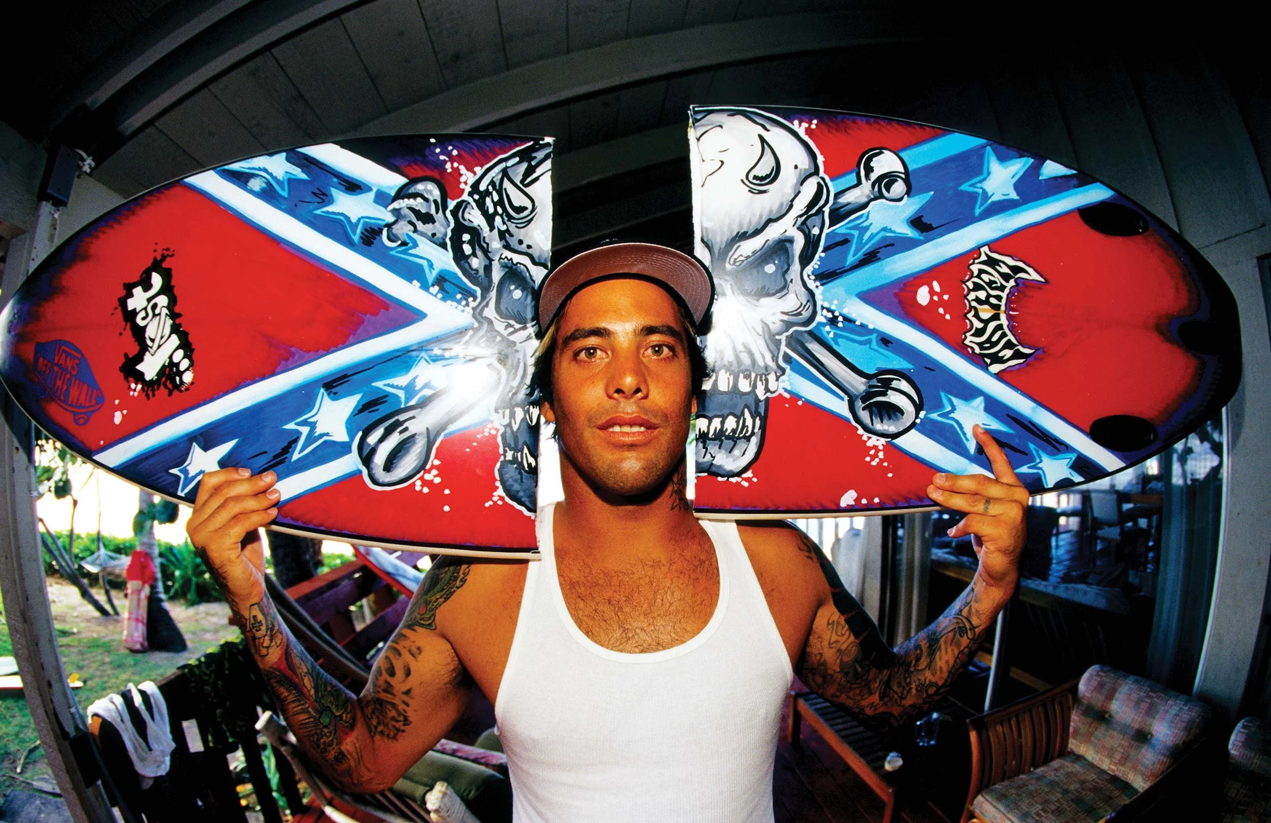 A person with tattoos wearing a white tank top and a cap is holding a skateboard with colorful artwork featuring skulls, stars, and graffiti. The person is standing outdoors in a shaded area.