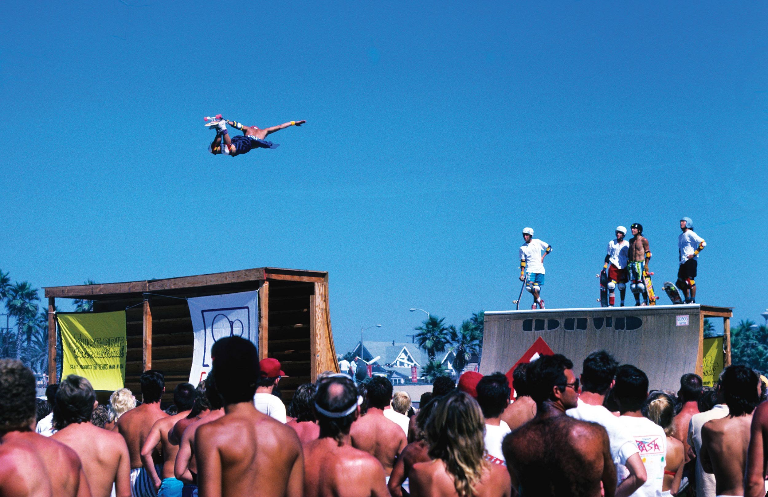 A crowd of shirtless people watching a skateboard stunt show outdoors on a sunny day, with a person mid-air doing a trick on a ramp and four others standing on the ramp in helmets and skateboards.