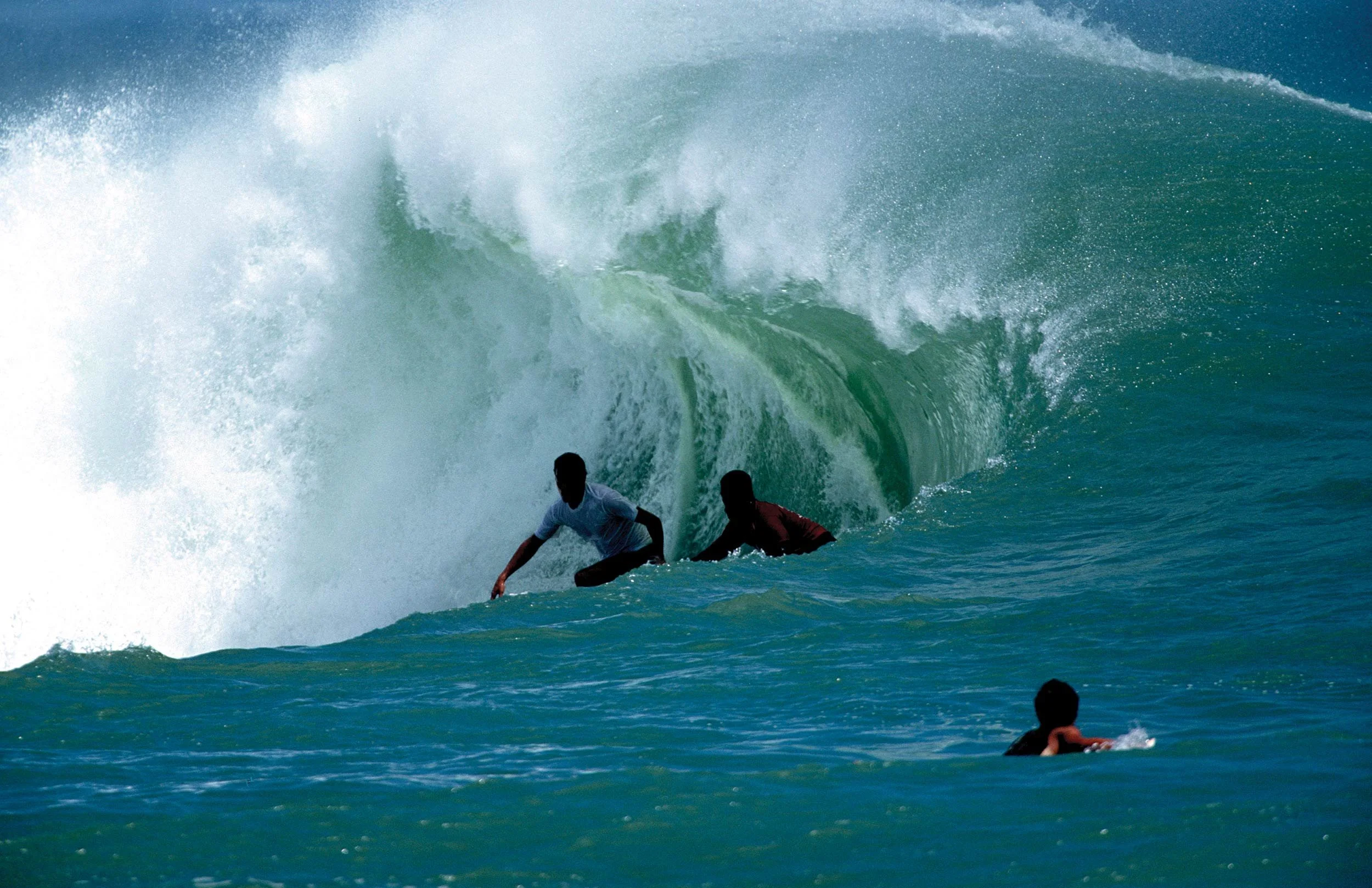Two surfers riding a large wave in the ocean with other people swimming nearby.