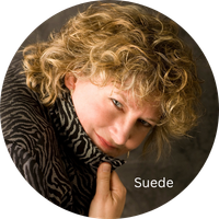 Close-up of a woman with curly blonde hair wearing a black patterned top and a scarf, looking at the camera with her hand near her chin.