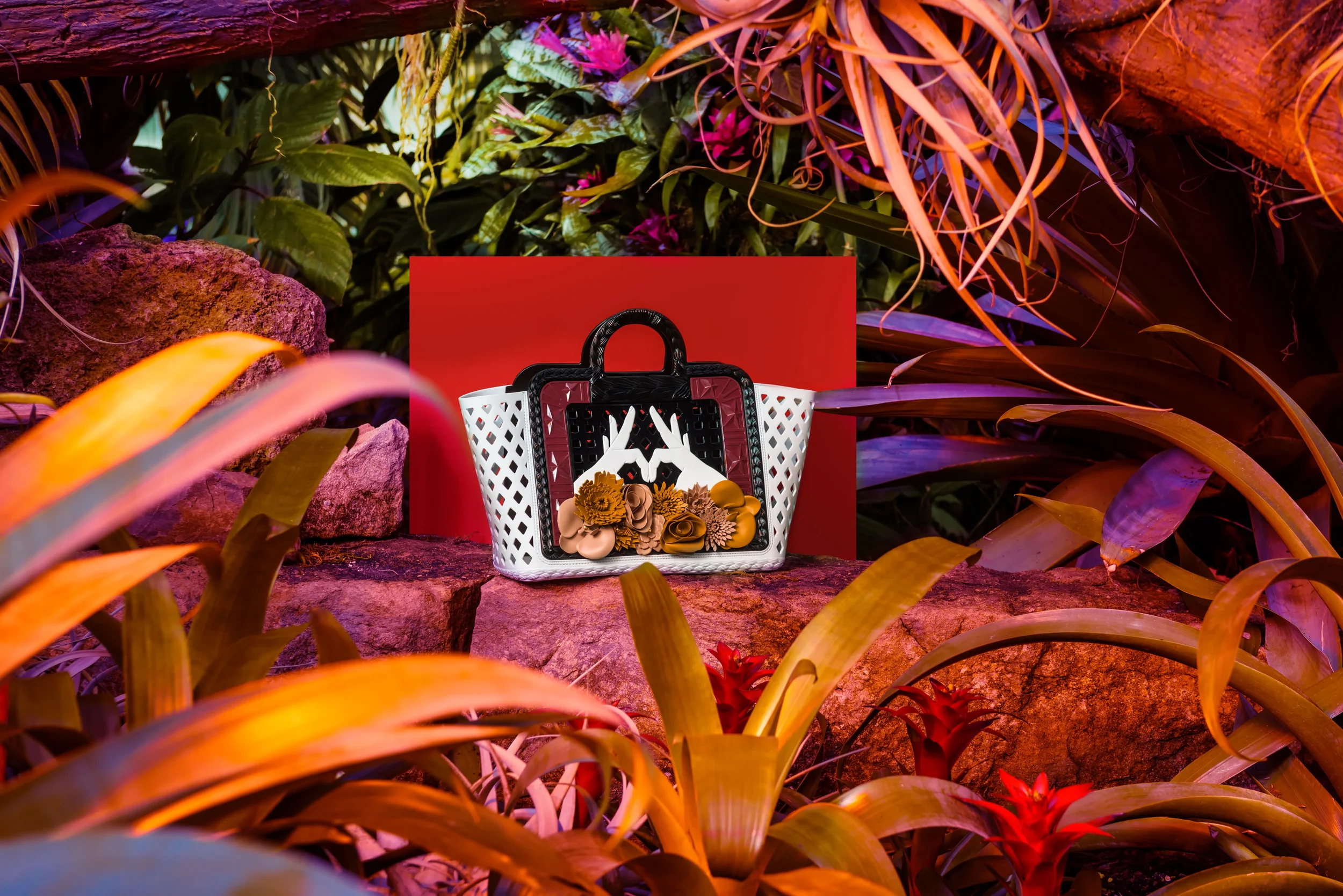 A designer bag with black, white, and red colors featuring floral decorations sits on a red surface, surrounded by tropical plants and rocks in a lush, colorful jungle setting with artificial lighting.