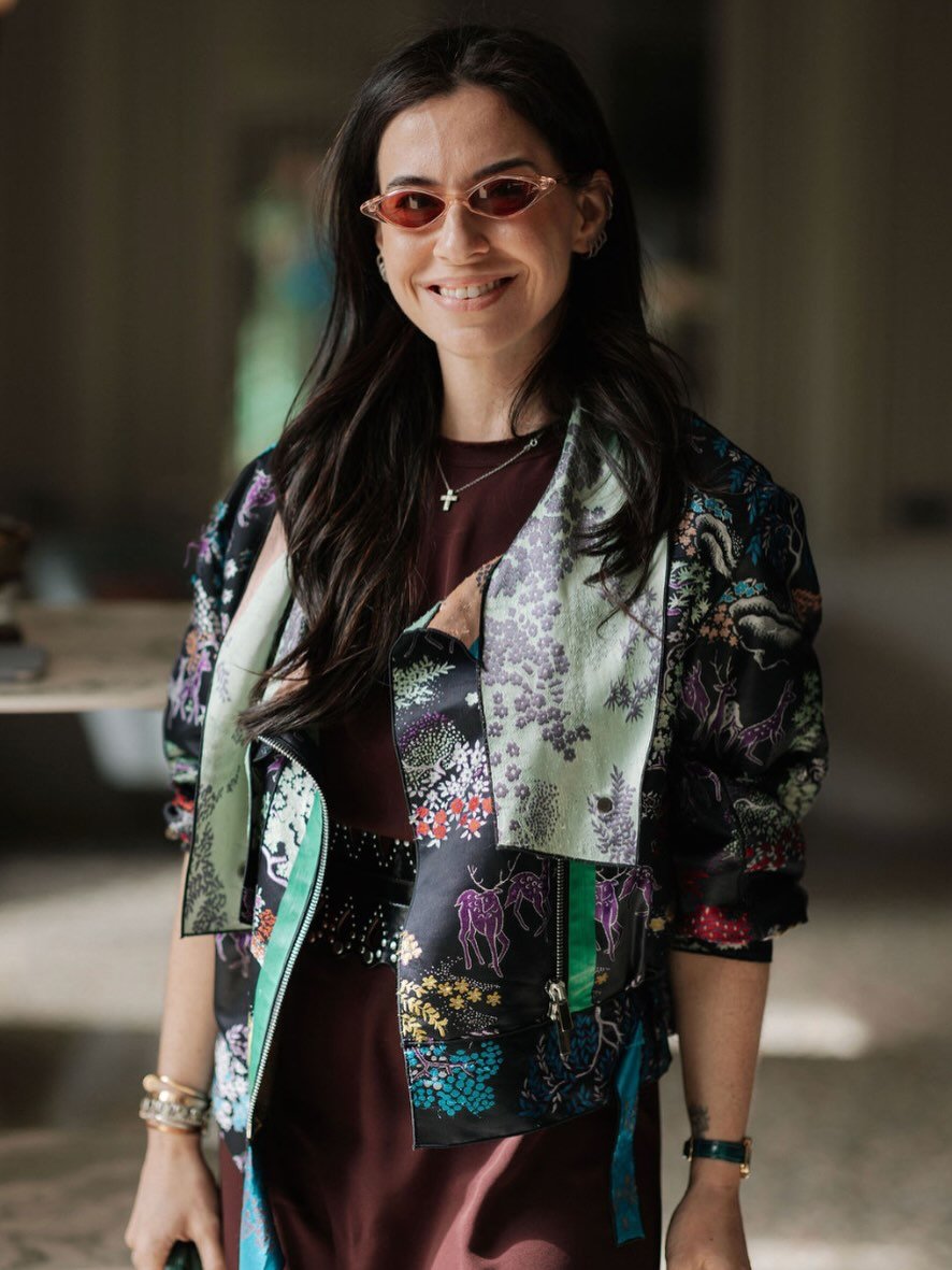 A smiling woman with long dark hair wearing pink-tinted sunglasses, a botanical print bomber jacket, a maroon dress, and jewelry, standing indoors.