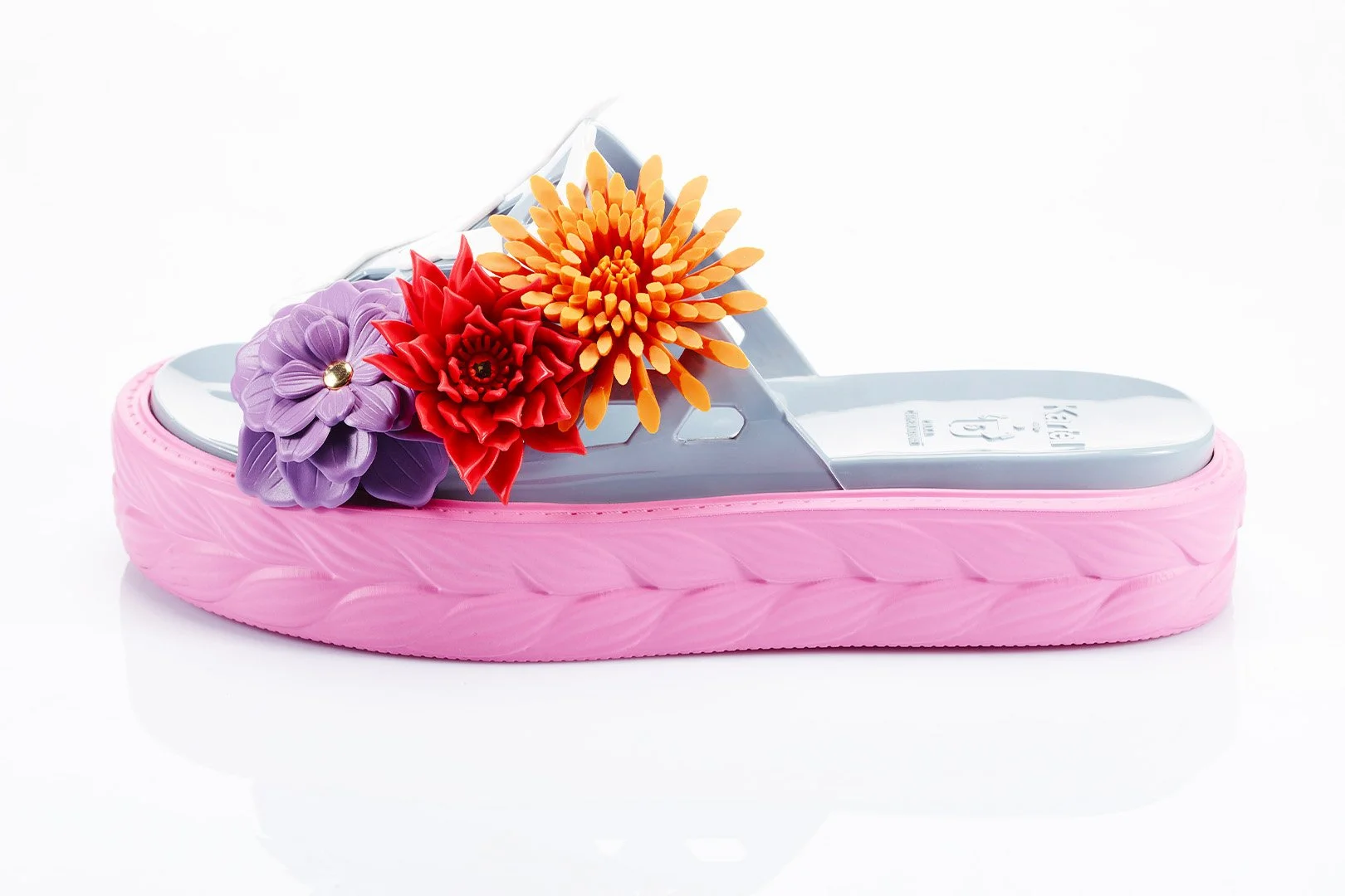 A pink platform sandal with a floral decoration on the strap, featuring purple, red, and orange artificial flowers against a white background.