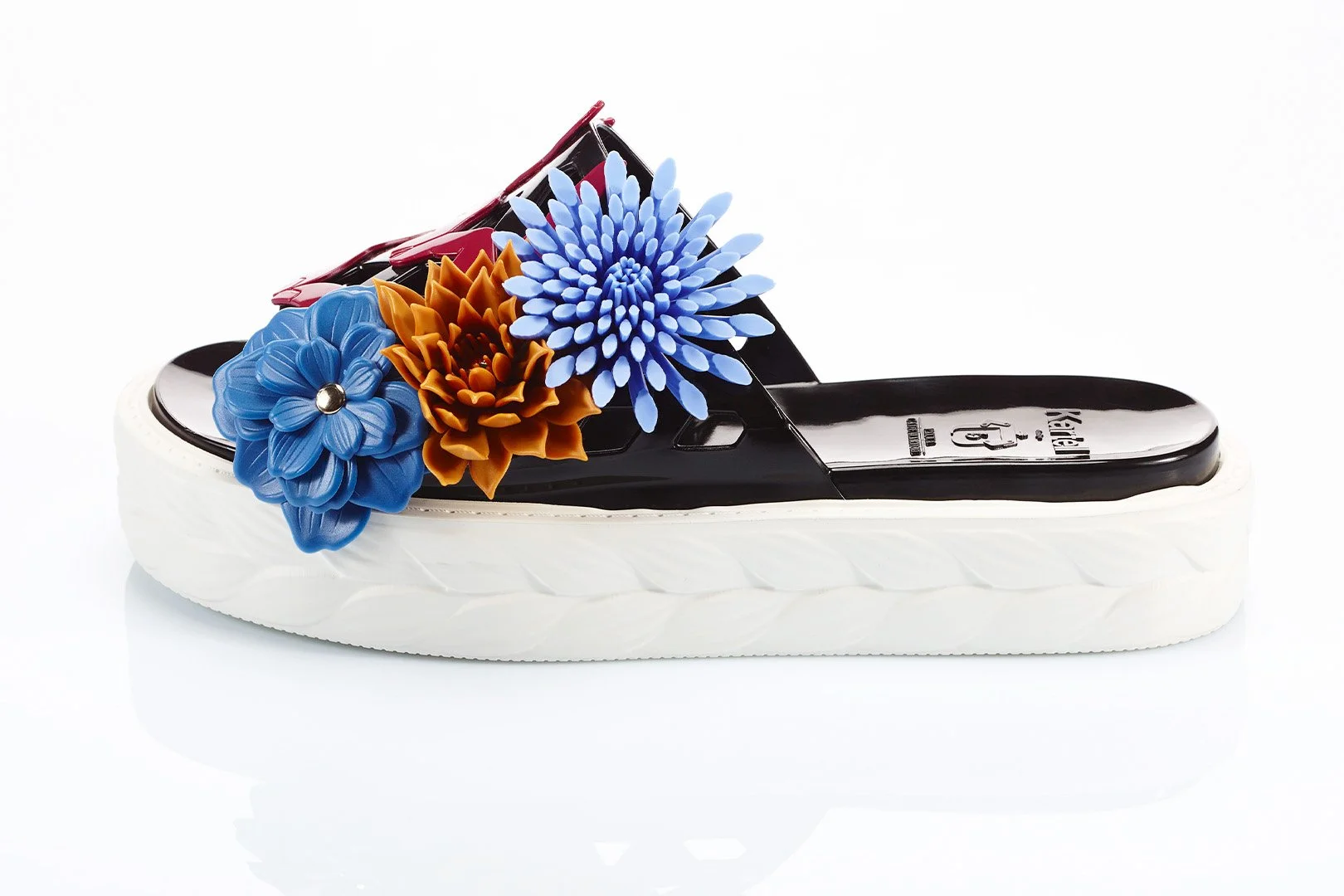 A black and white slide sandal with large colorful plastic flowers on the strap, featuring blue, orange, and purple flowers.