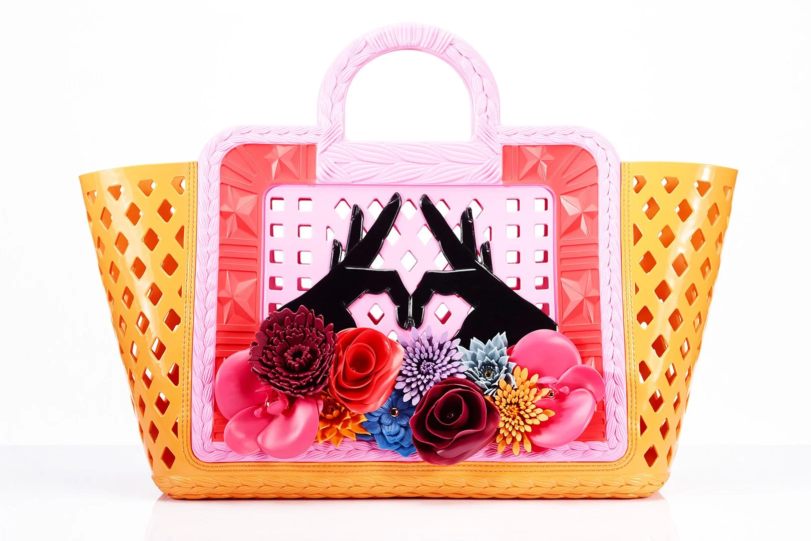 A colorful handbag with a pink handle, yellow side panels, and a pink front panel decorated with black hands making a heart shape, surrounded by multicolored flowers.