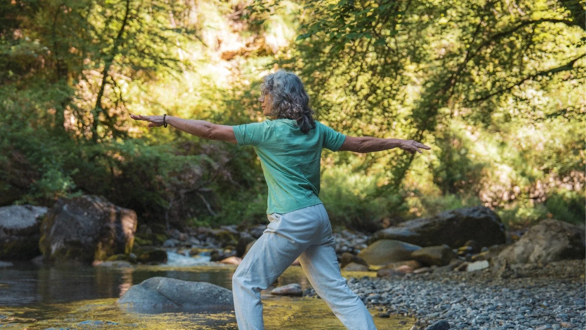 Une femme âgée pratiquant le yoga ou la méditation dans une forêt en marchant dans un ruisseau, entourée d'arbres verts et d'une lumière douce.