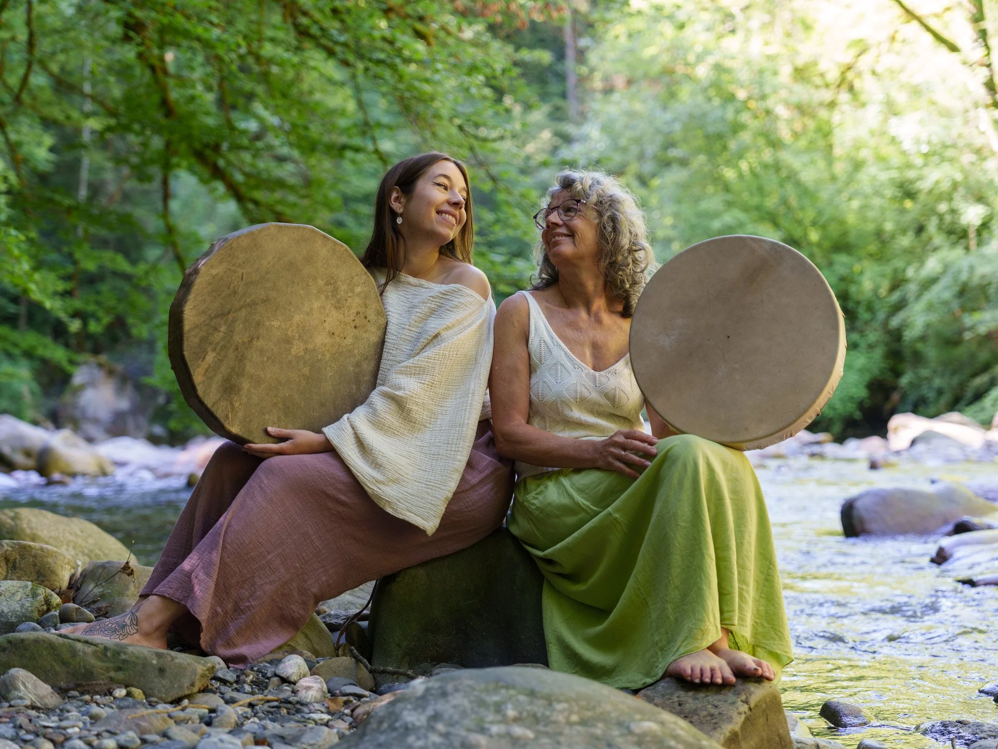 Deux femmes assises sur une roche dans un ruisseau, tenant des tambours en cuir, entourées de nature avec des arbres verdoyants et une lumière douce.
