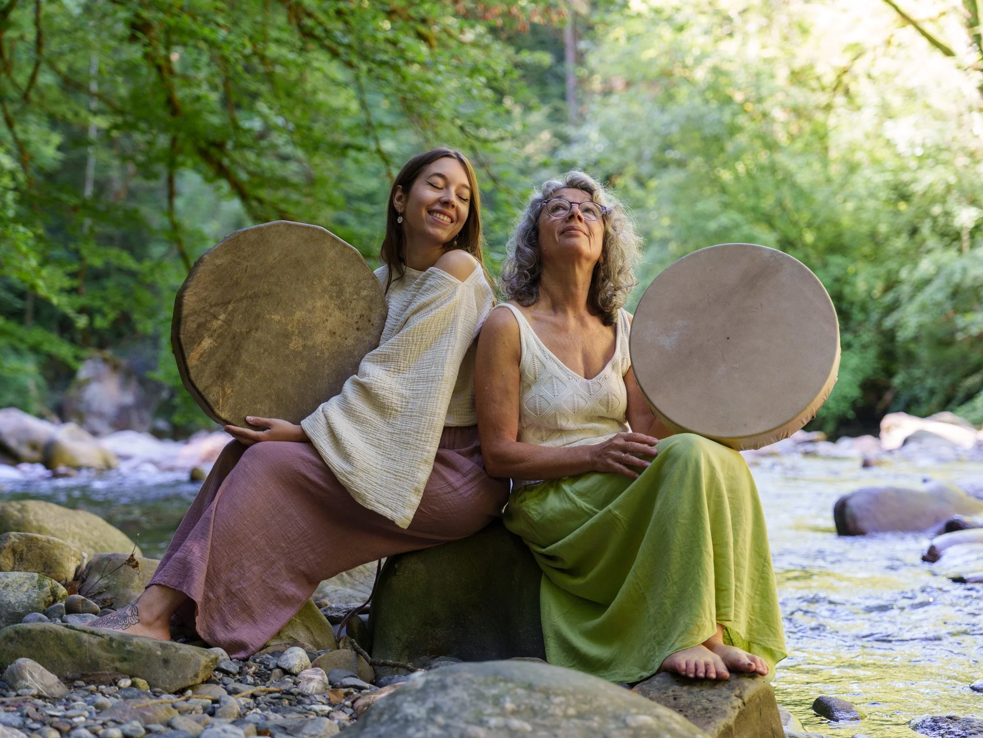 Deux femmes assises sur un rocher au bord d'une rivière dans une forêt, tenant des tambours originaux, souriant et profitant d'une journée en plein air.
