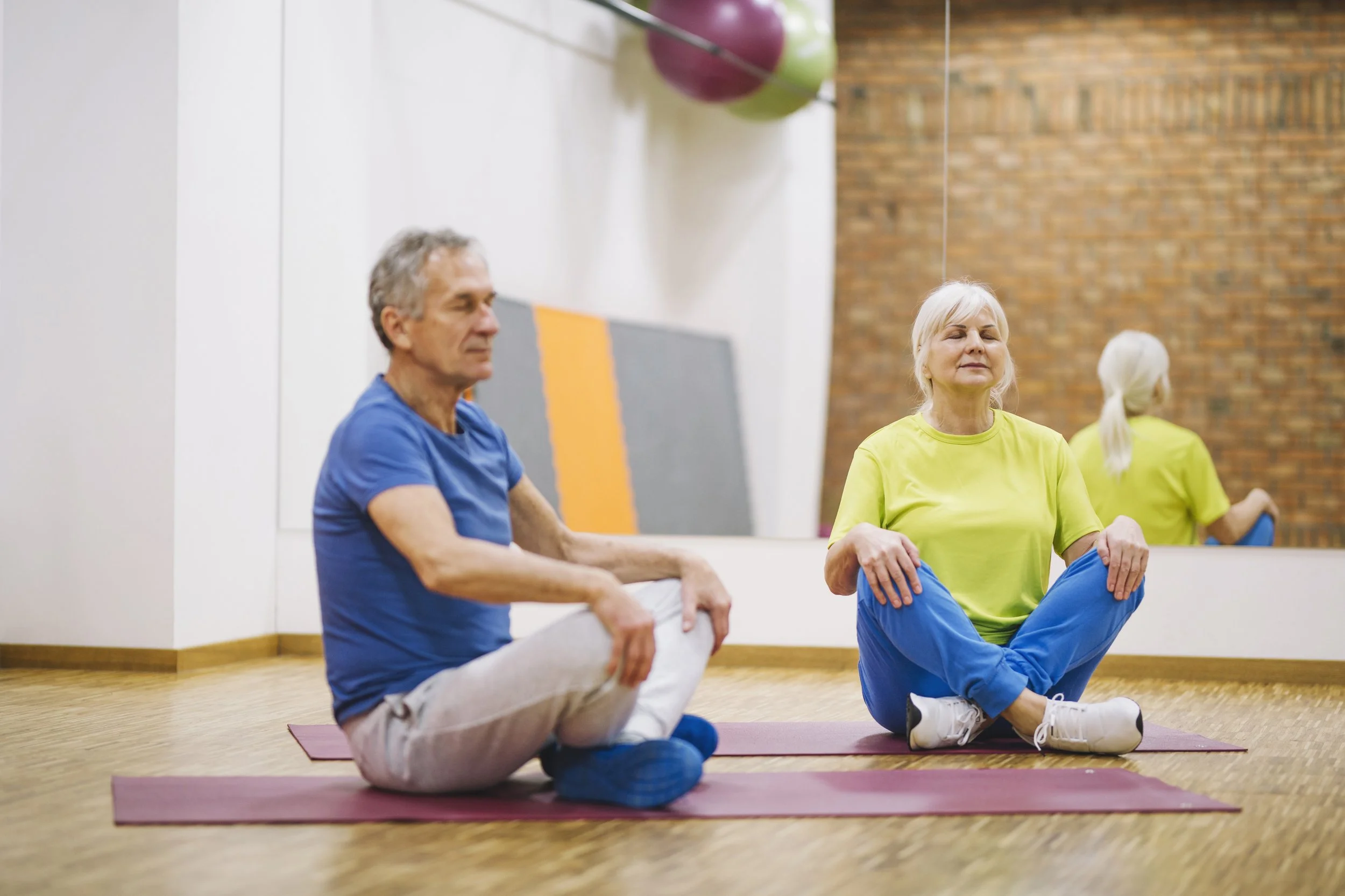 Deux personnes âgées, un homme et une femme, pratiquant la méditation assis sur un tapis de yoga dans une salle de sport. Ils portent des vêtements de sport colorés et semblent concentrés et détendus.