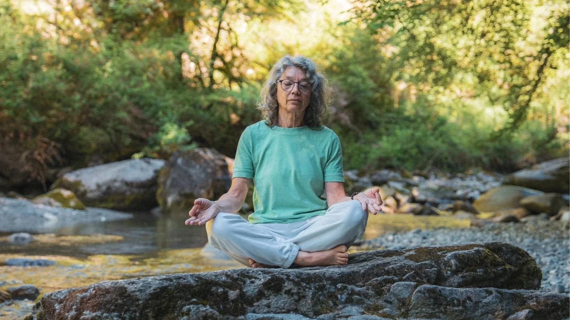 Une personne âgée pratiquant la méditation en position lotus près d'une rivière dans un environnement forestier en automne.