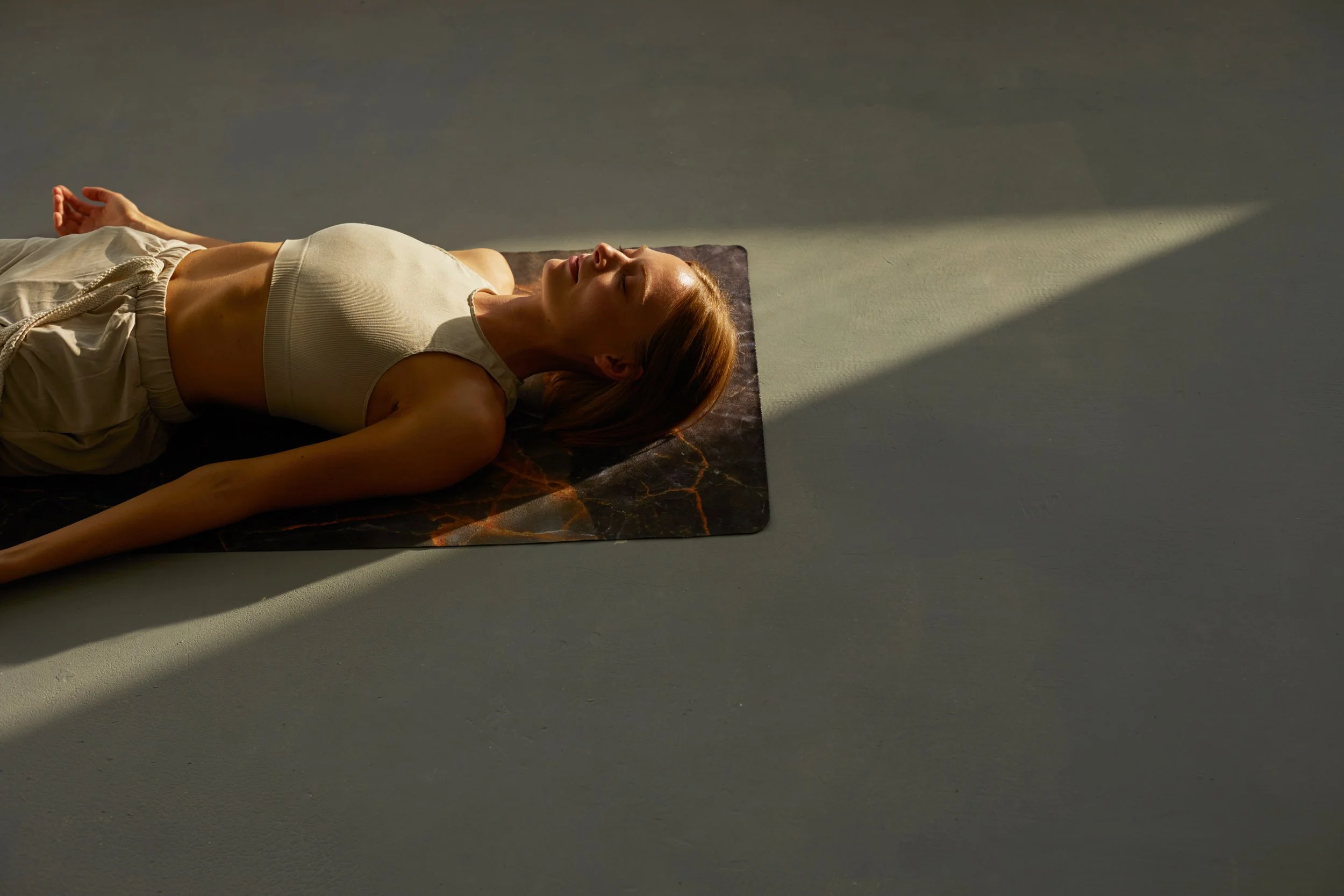 Une femme allongée sur un tapis de yoga, dans une pièce calme, profitant d'un moment de relaxation avec la lumière du soleil qui filtre à travers la fenêtre.