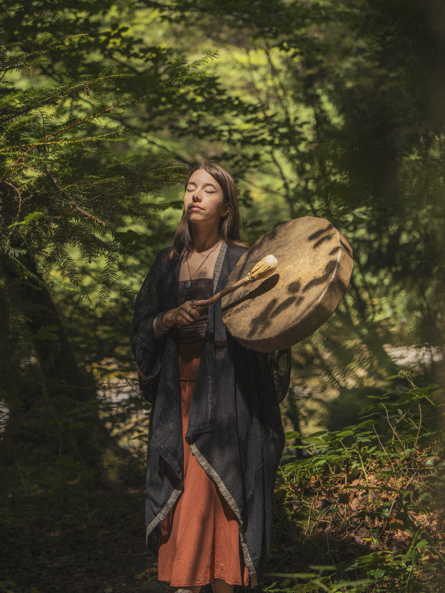 Une jeune femme méditant dans une forêt, avec les yeux fermés et un bâton rond dans les mains, entourée de verdure et de feuillage dense.