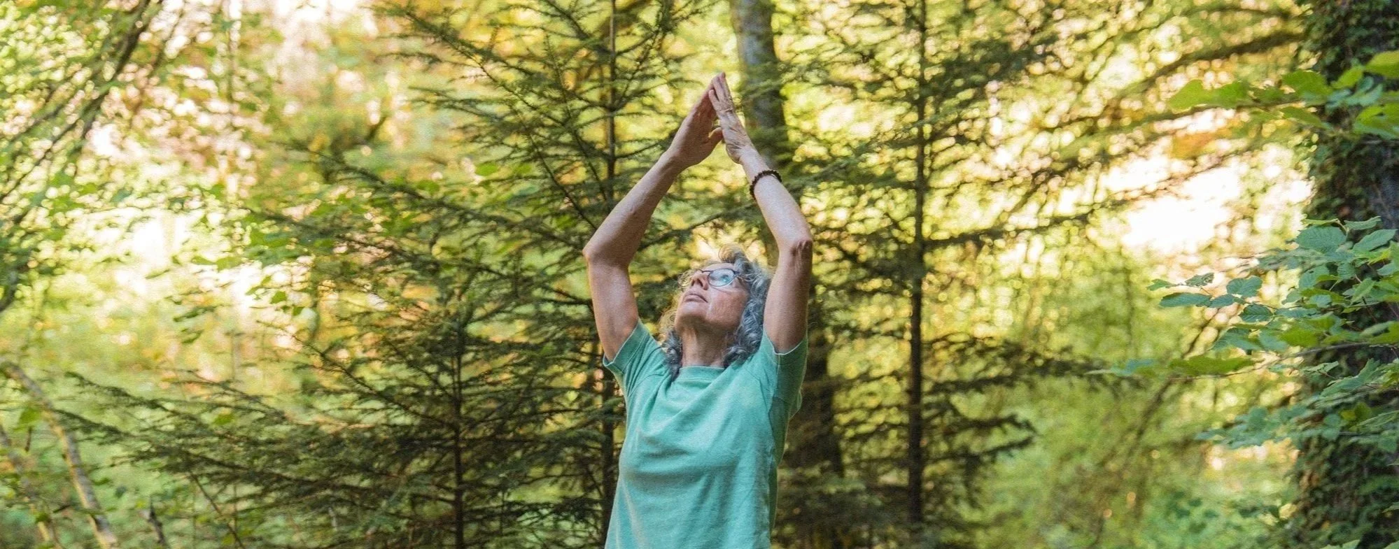 Une femme âgée, portant des lunettes et un t-shirt vert, pratique le yoga ou la méditation dans une forêt en automne, avec des mains levées au-dessus de la tête et formant une pyramide, entourée d'arbres aux feuilles vertes et dorées.
