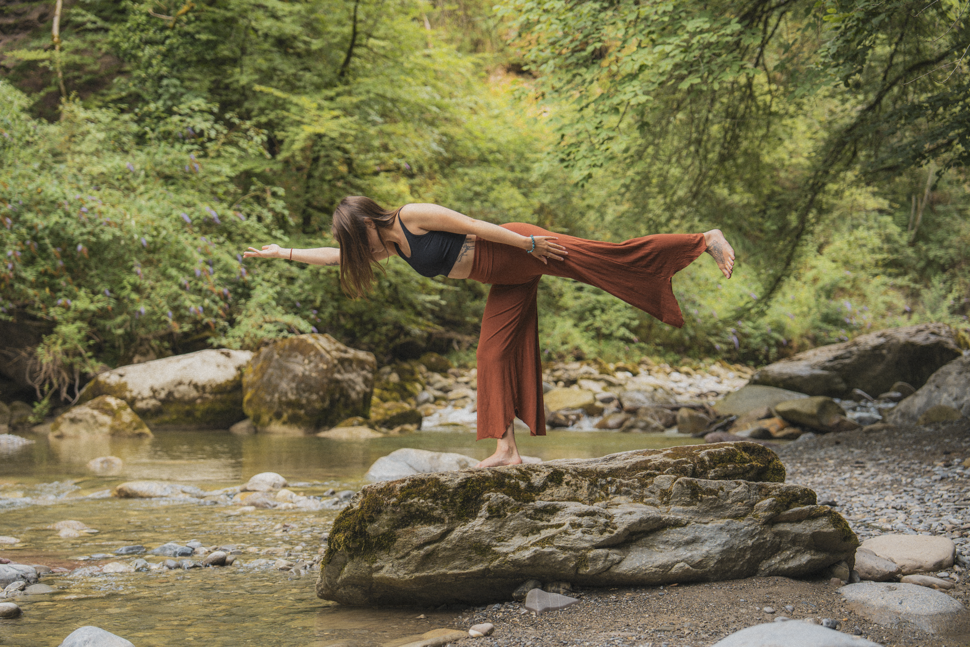 Une femme effectue une pose d'équilibre dans la nature, au bord d'une rivière, entourée de rochers et d'une forêt dense et verdoyante.