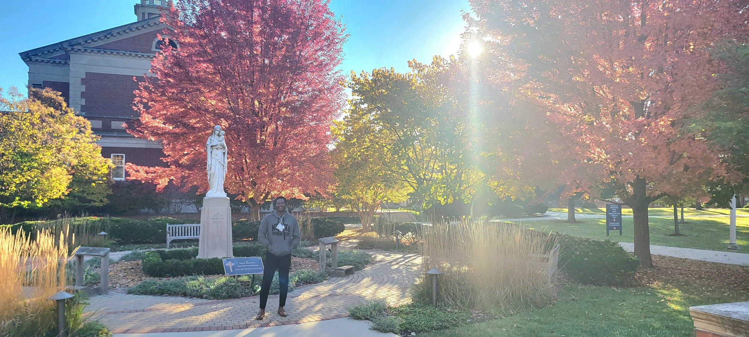 A person standing in a garden with a statue of the Virgin Mary, surrounded by colorful autumn trees, with sunlight streaming through the branches.