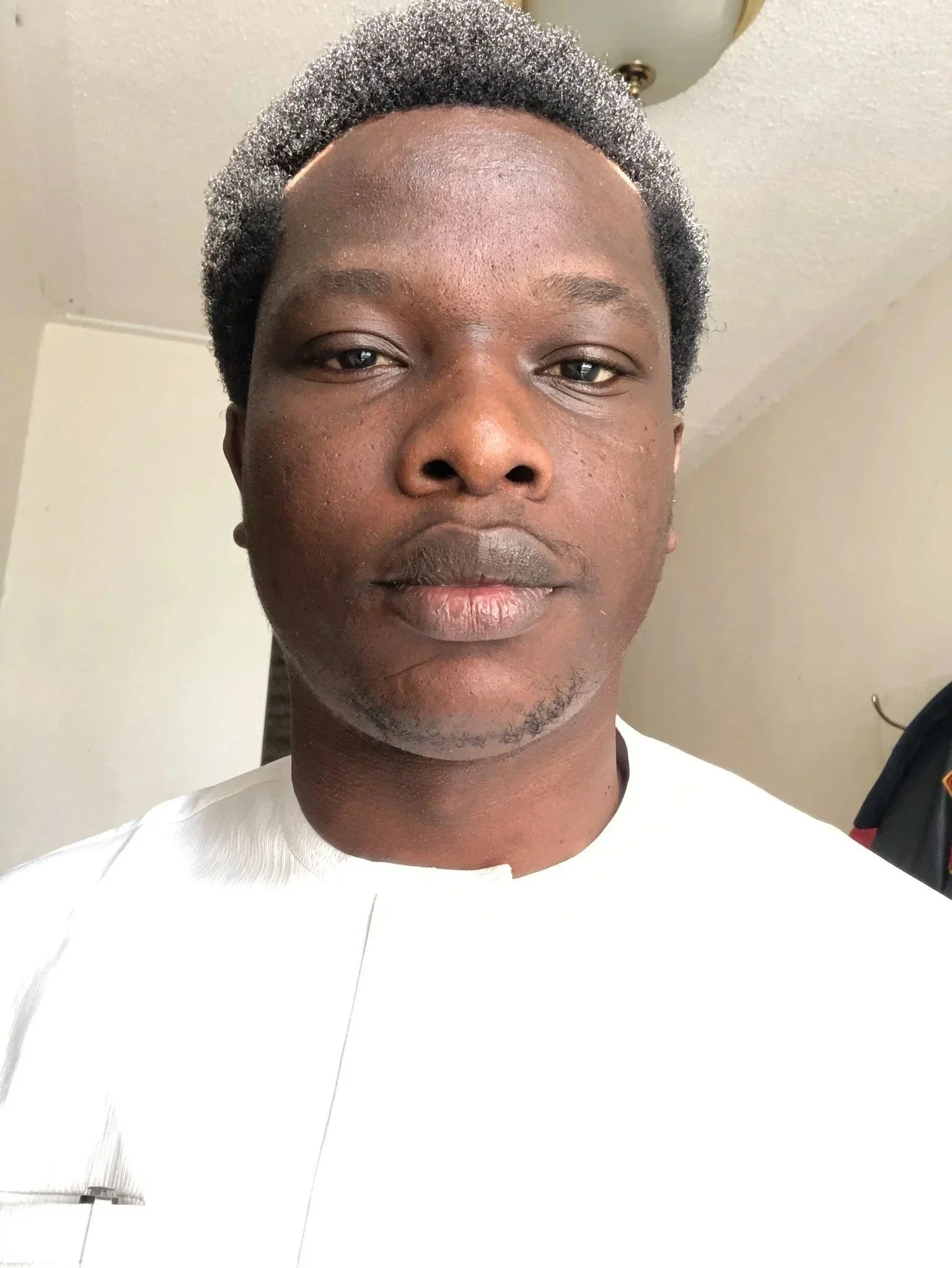 Close-up of a person with short gray hair wearing a white shirt, standing indoors.