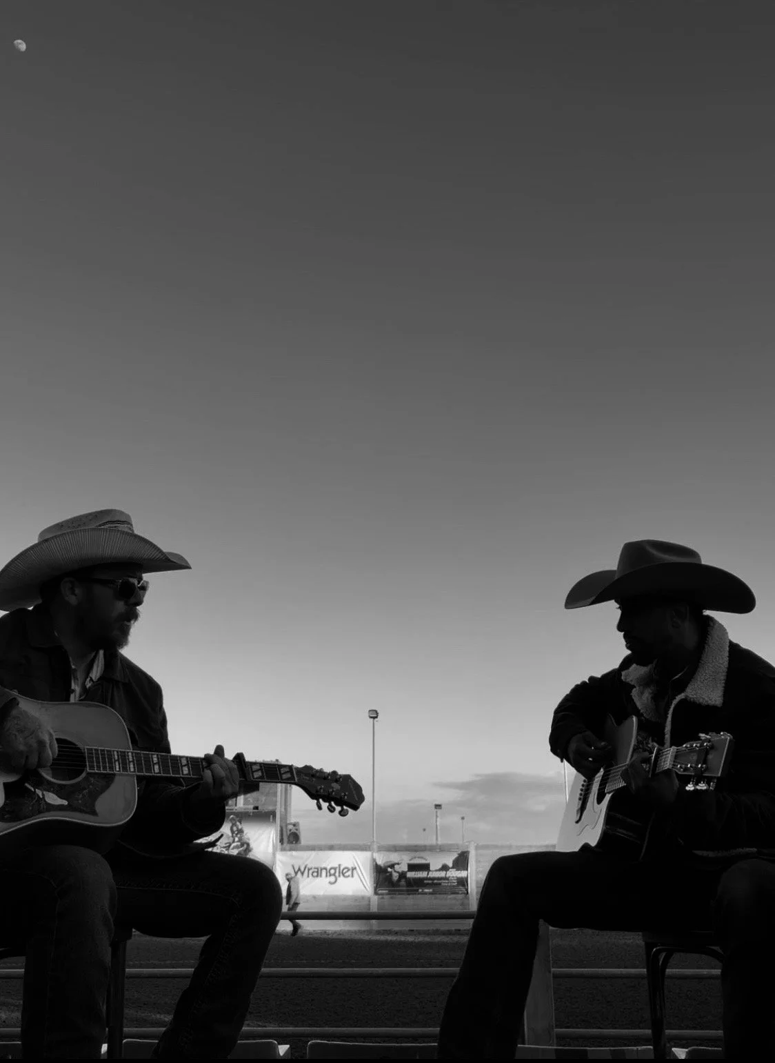 Two men wearing cowboy hats and sunglasses playing acoustic guitars outdoors during sunset.
