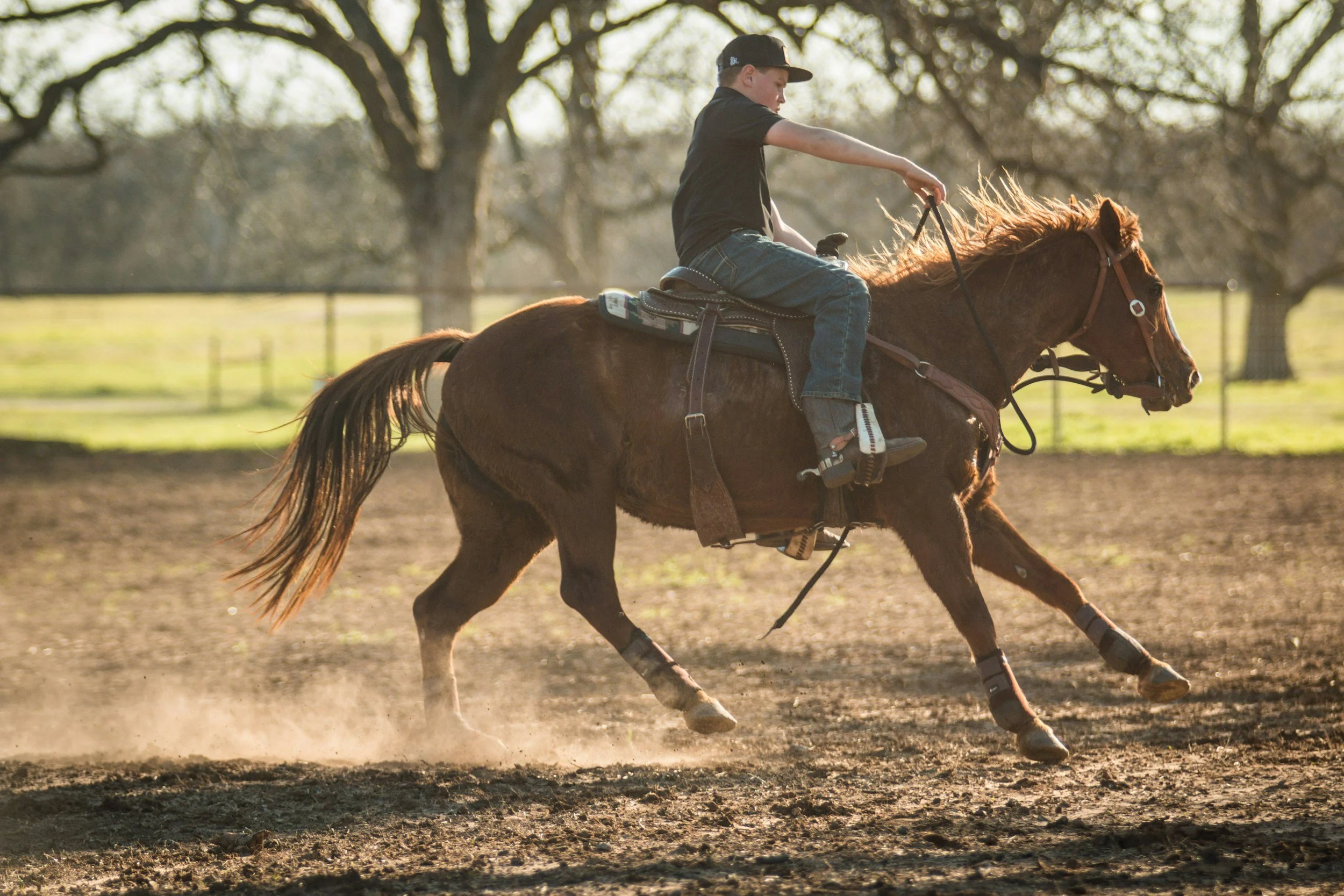 A young man riding a brown horse on a dirt field with trees in the background during daytime.