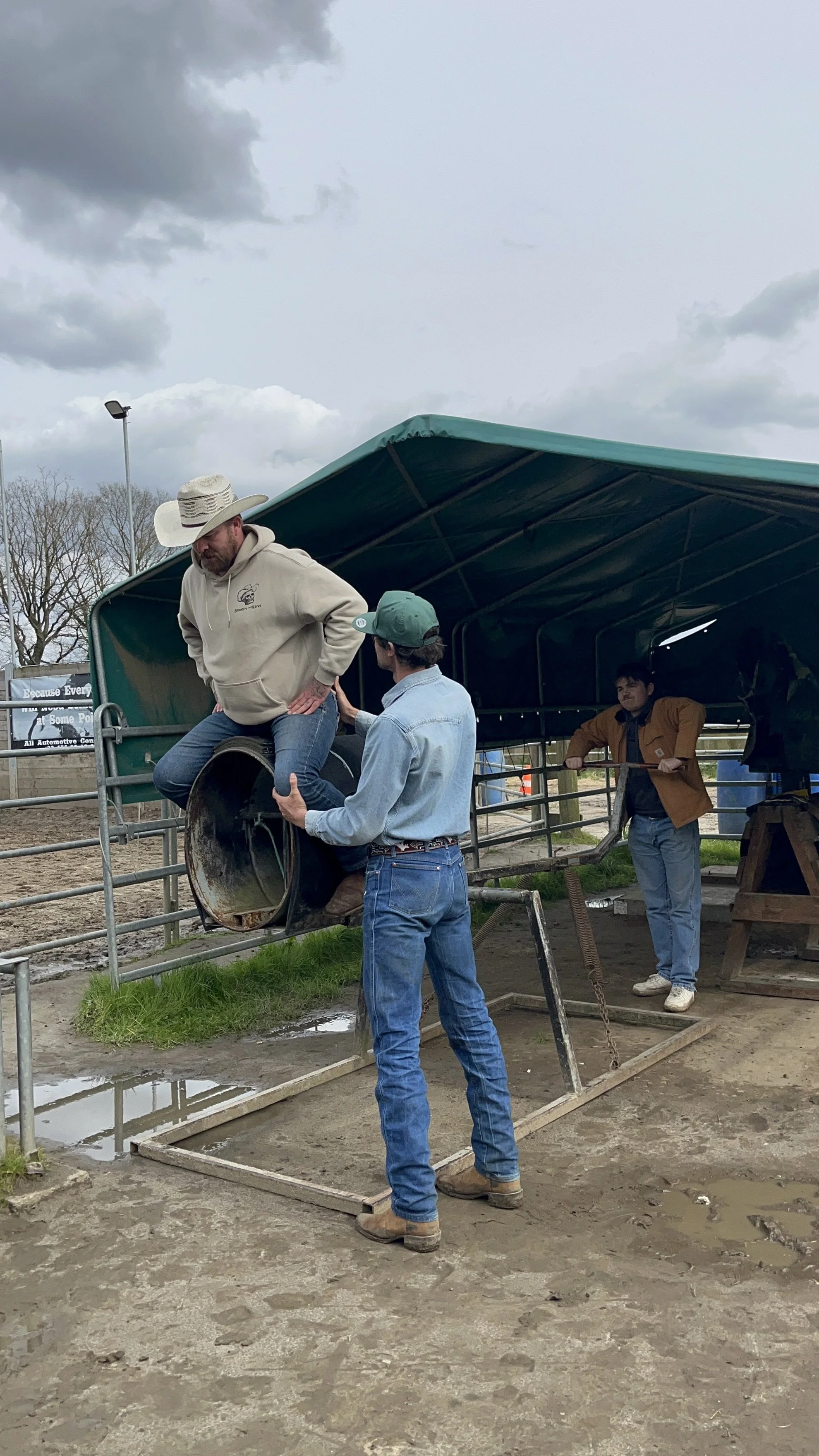 Three men at a fairground are involved in a rodeo activity. One man is sitting in a metal barrel or rodeo saddle, with another man assisting him. The third man is standing nearby, holding the structure. The setting is outdoors with cloudy skies and a