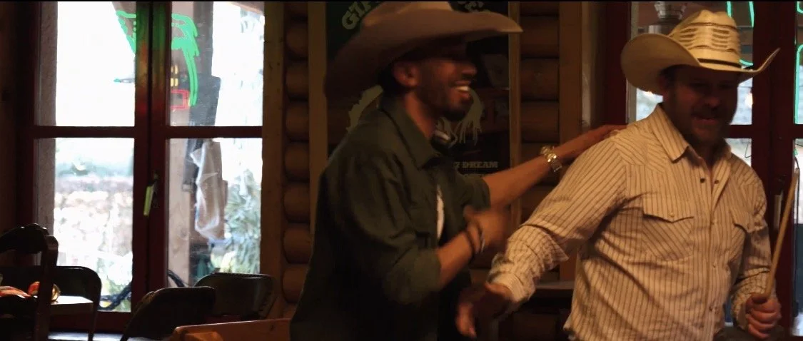 Two men wearing cowboy hats inside a log cabin-style restaurant, one patting the other's shoulder.