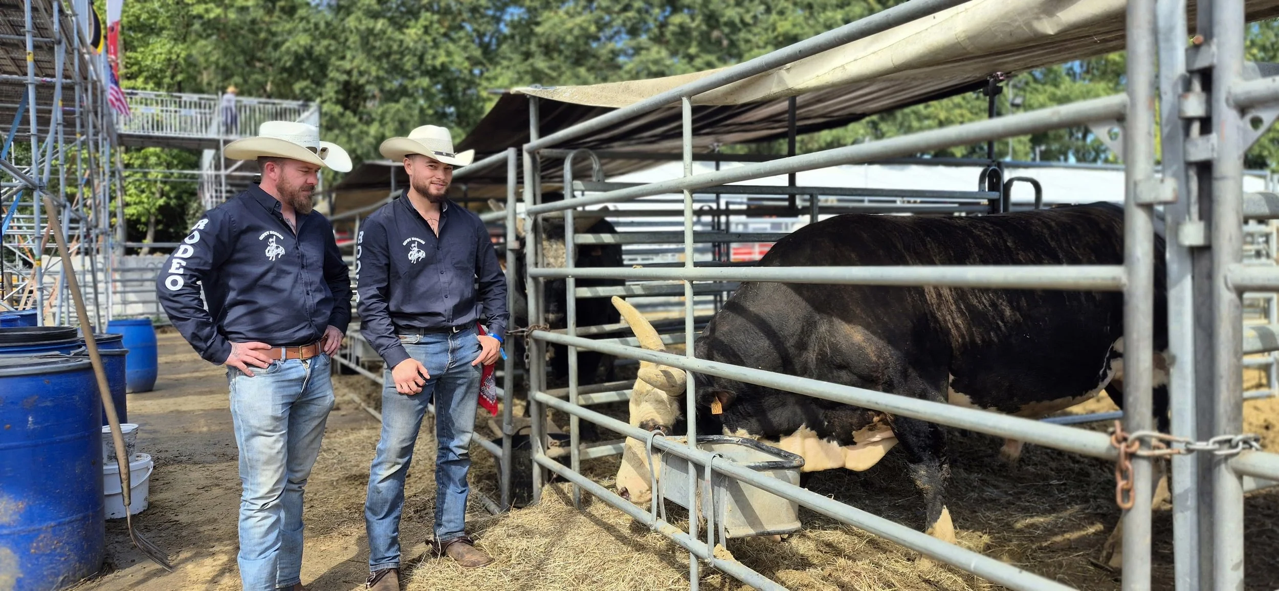 Two men wearing cowboy hats and navy shirts observing a black and white cow in a cattle pen at an outdoor event.