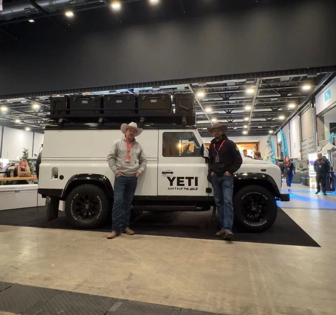 Two men in cowboy hats standing next to a white YETI off-road vehicle with a large roof cargo box at an indoor event.