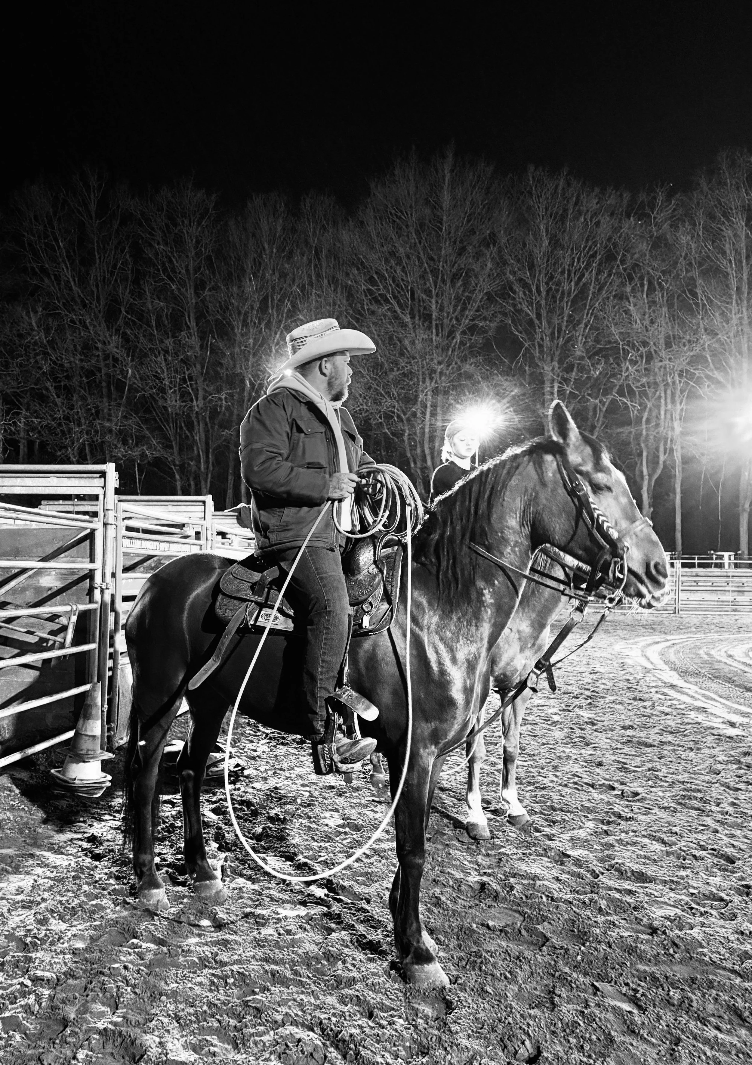 A man wearing a cowboy hat and jacket sitting on a horse with a young girl in the background, at night in an outdoor arena with bright lights and leafless trees.