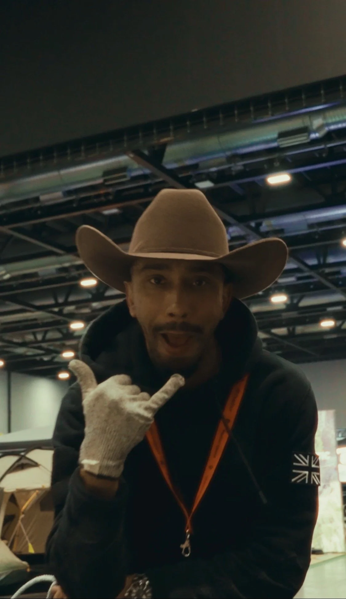 A man wearing a cowboy hat and a black hoodie with a Union Jack patch on the sleeve, making a shaka hand gesture and smiling at the camera.