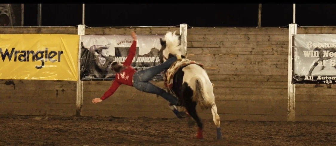 A rodeo rider falling off a bucking horse inside an arena with wooden walls and advertisements.