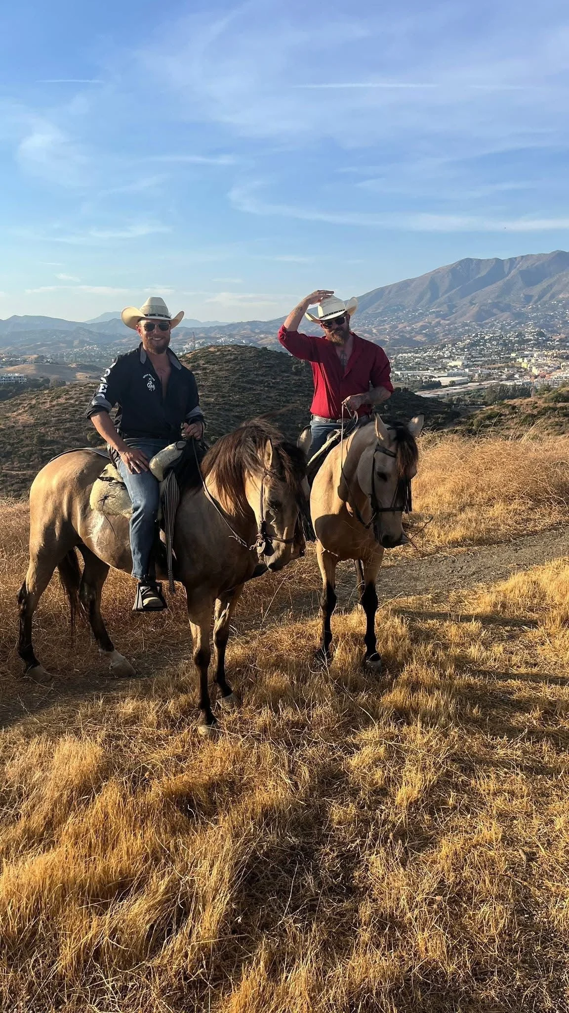 Two men riding horses on a dry, grassy hillside with a city and mountains in the background. One is wearing a cowboy hat and sunglasses, the other is tipping his hat.