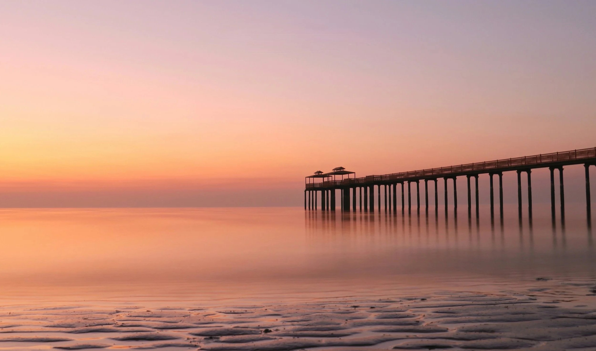 A pier extending into calm water during sunset, with umbrellas on the pier and a pastel-colored sky reflecting on the water.