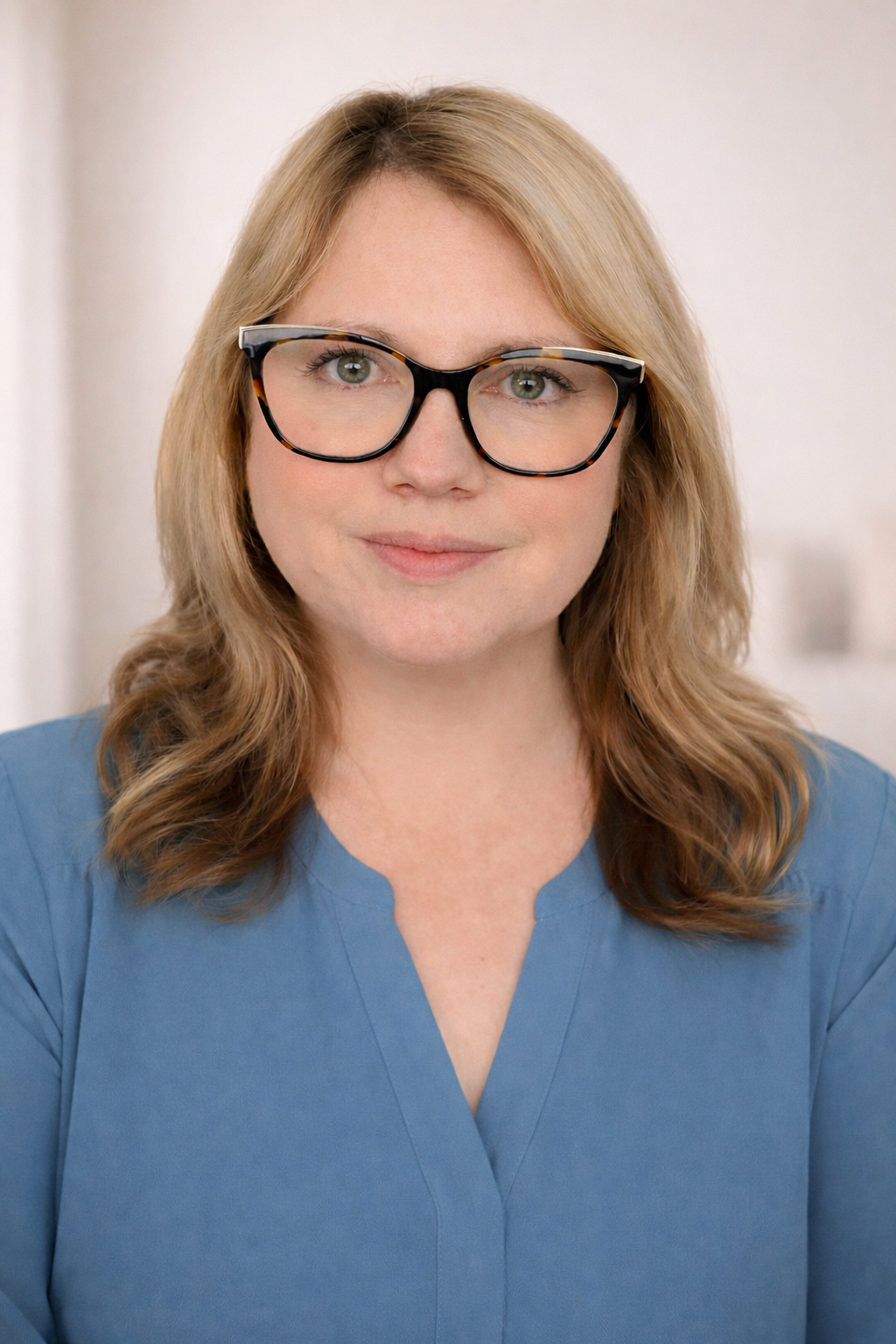 A woman with shoulder-length wavy blonde hair wearing large black-rimmed glasses and a blue top, smiling softly in a bright, neutral indoor setting.