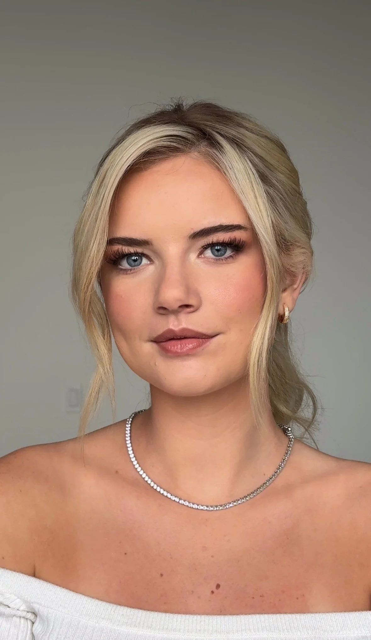 A young woman with blonde hair styled in loose waves, blue eyes, and light makeup, wearing a white off-the-shoulder top and jewelry including a diamond necklace, small hoop earrings, and a ring, standing against a plain light-colored background.