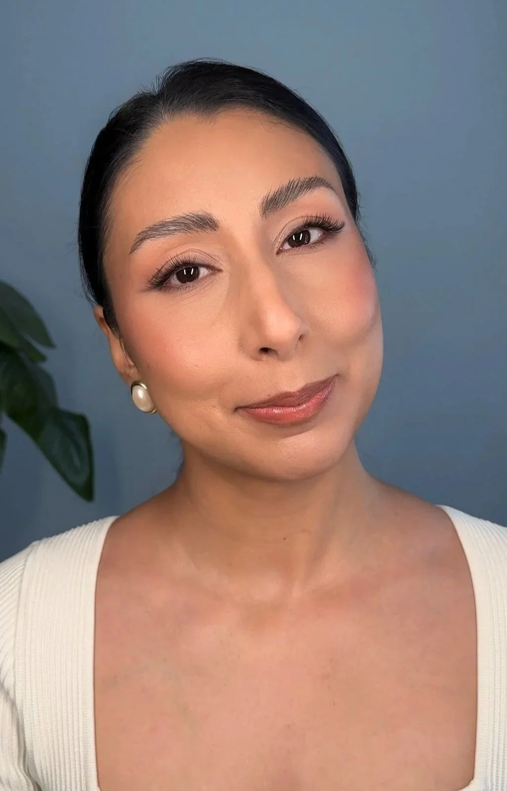Close-up of a woman with dark hair, makeup, pearl earrings, wearing a white top, standing against a blue background with part of a green plant visible. Wedding Makeup. Bridal Makeup.