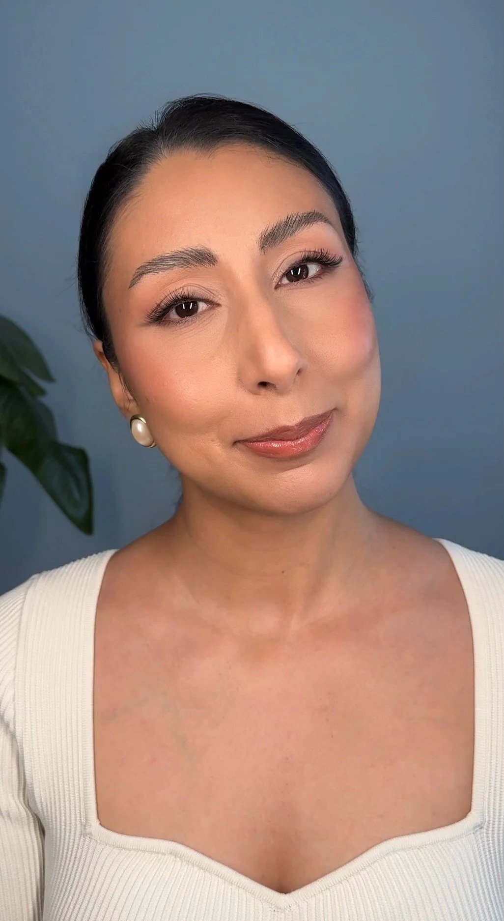 A woman with dark hair styled back, soft makeup, pearl earrings, wearing a white ribbed top with a square neckline, smiling gently in front of a blue background and a leafy plant. Bridal Makeup Look.