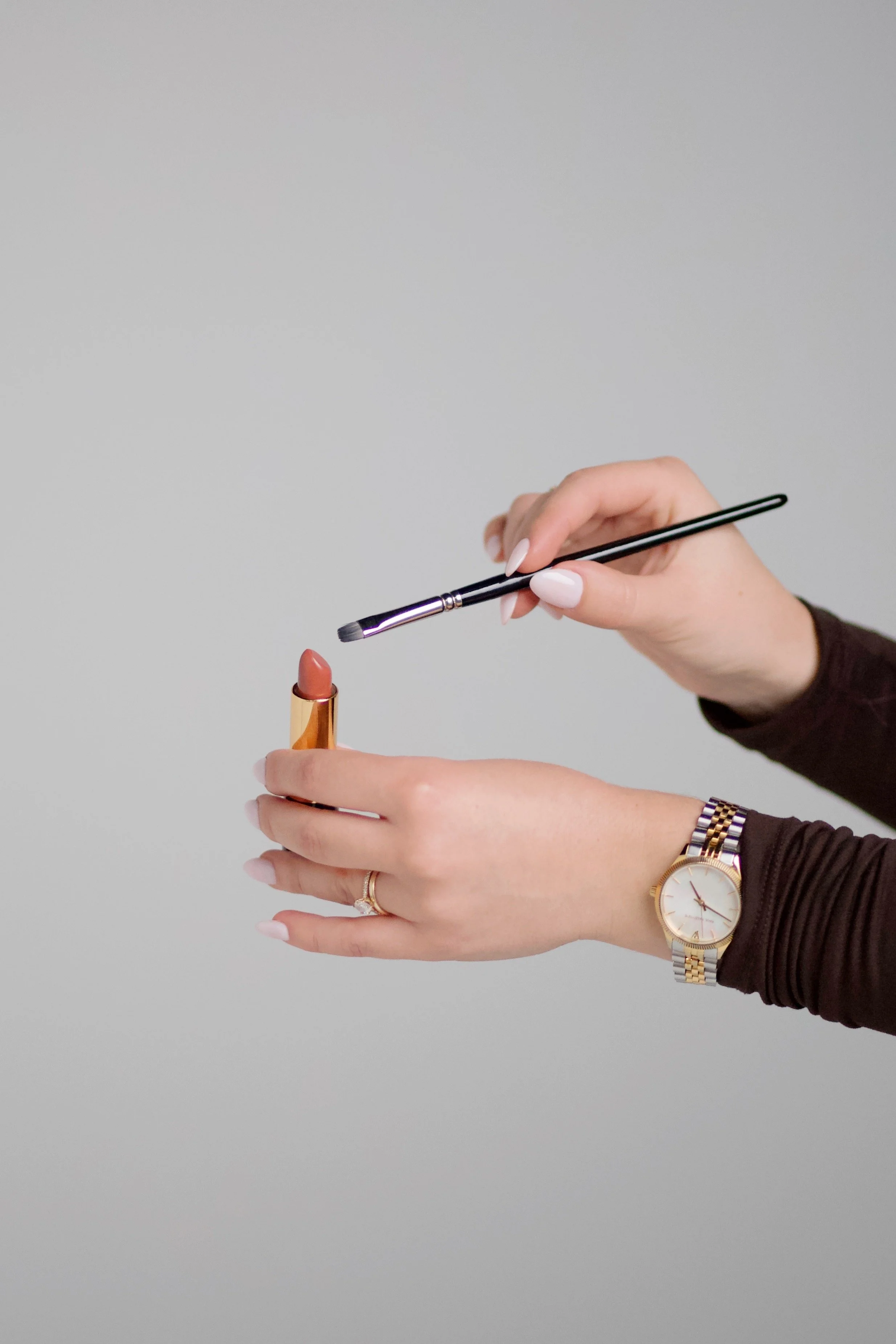 Person applying lipstick with a makeup brush, wearing a wristwatch and rings, against a plain light gray background. Makeup Artist using lipstick.
