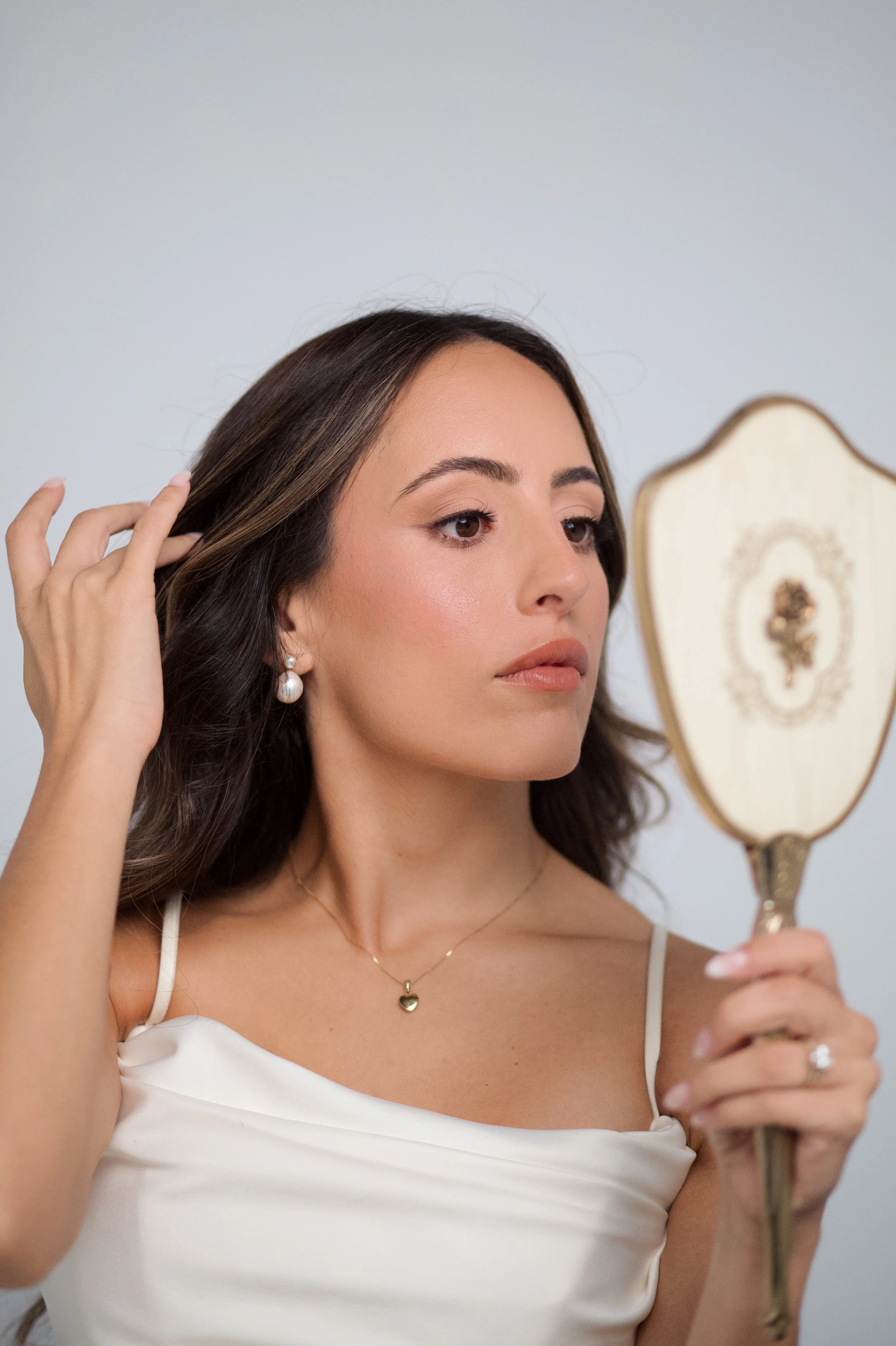 A woman with dark hair, wearing pearl earrings and a heart-shaped necklace, is looking at herself in a vintage hand mirror while adjusting her hair.
