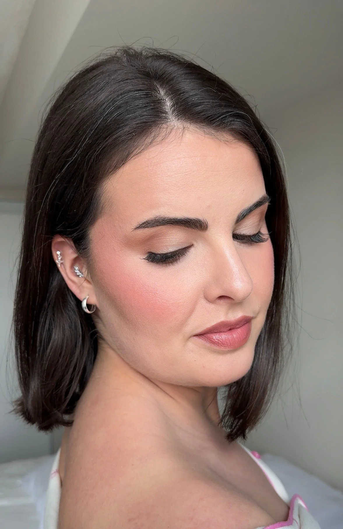 A close-up photo of a woman with dark brown hair, wearing earrings, with her eyes closed and head slightly tilted. She has makeup including foundation, blush, lipstick, eyeliner, and mascara. Bridal Makeup. Wedding Makeup. 