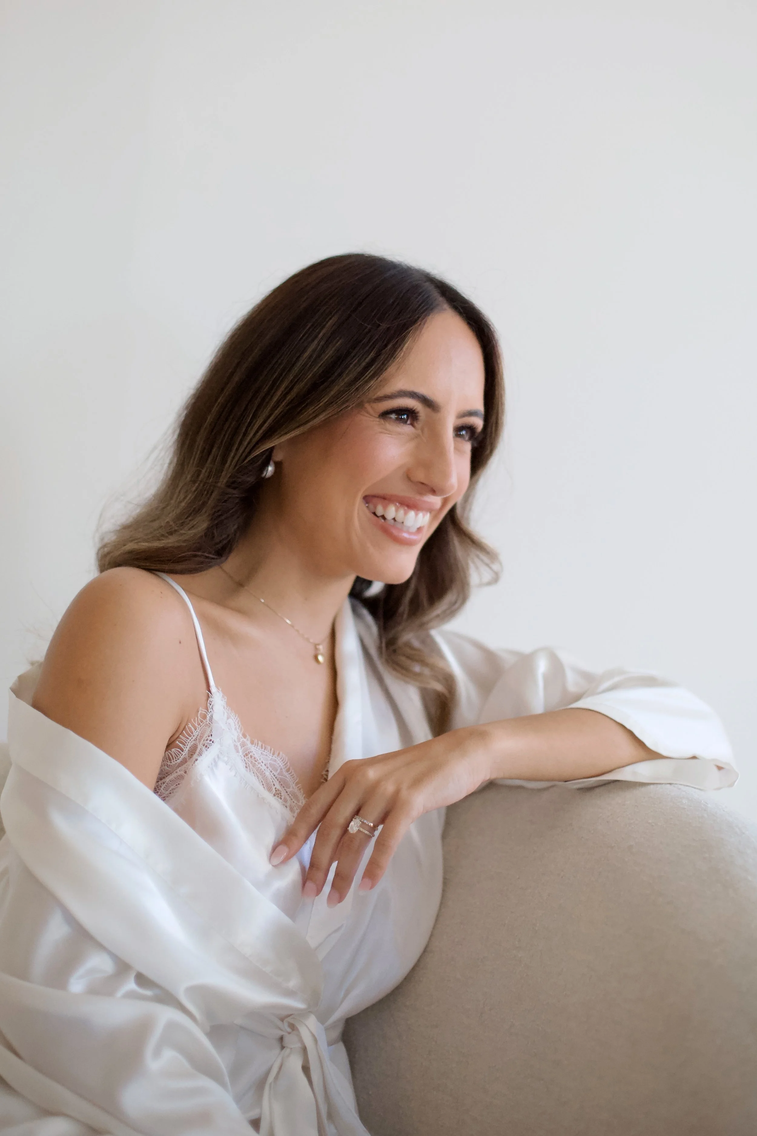A woman with long brown hair smiling, wearing a white satin robe with lace detail, sitting on a beige sofa with her arm resting on it, against a plain white background. Bride getting ready on her wedding morning. Happy Bride with beautiful Makeup.