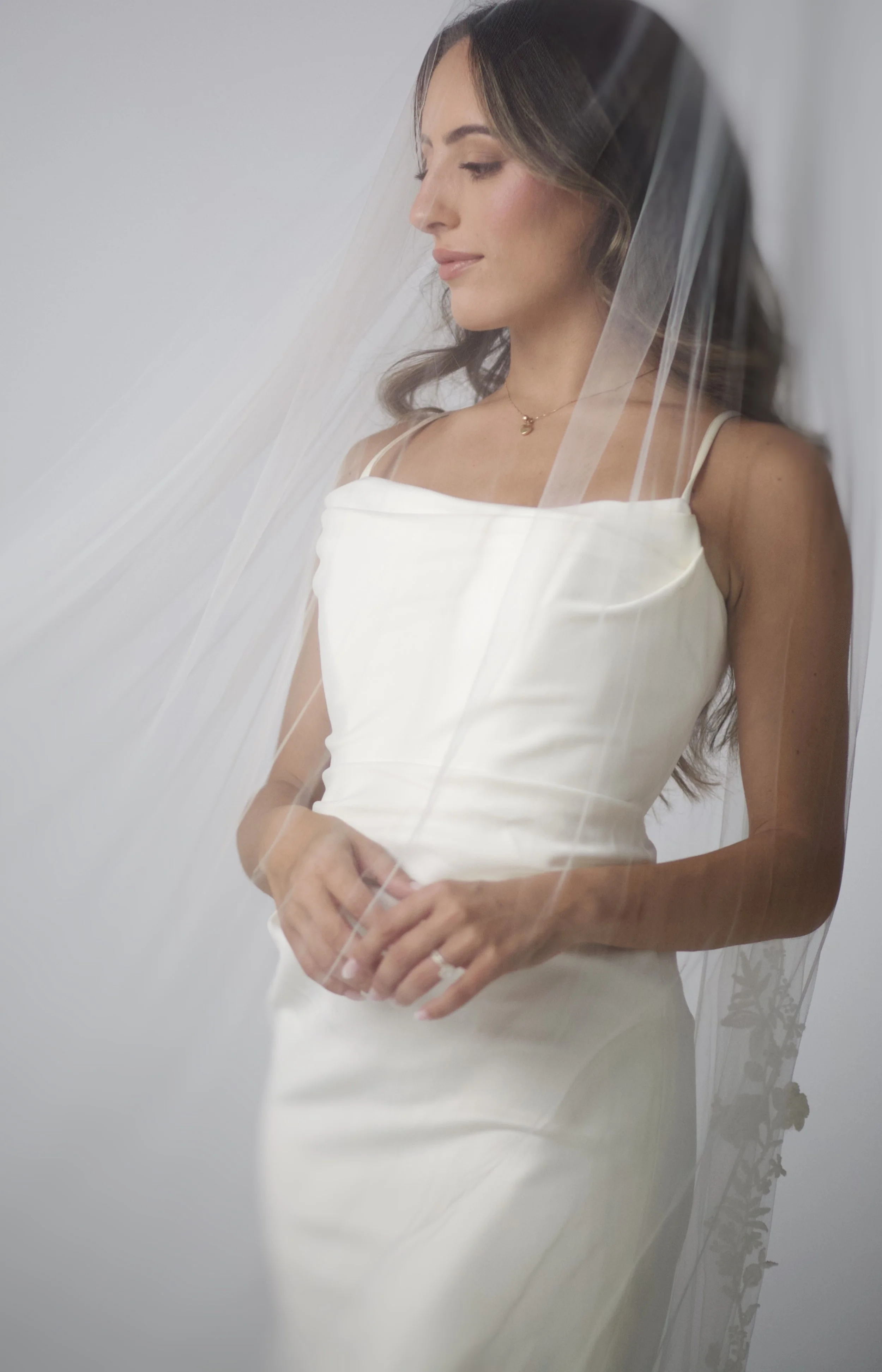 A woman in an elegant white wedding dress, standing with her hands clasped, wearing a wedding veil and a delicate necklace, looking slightly downward with a gentle expression. Bride. Toronto Bride.