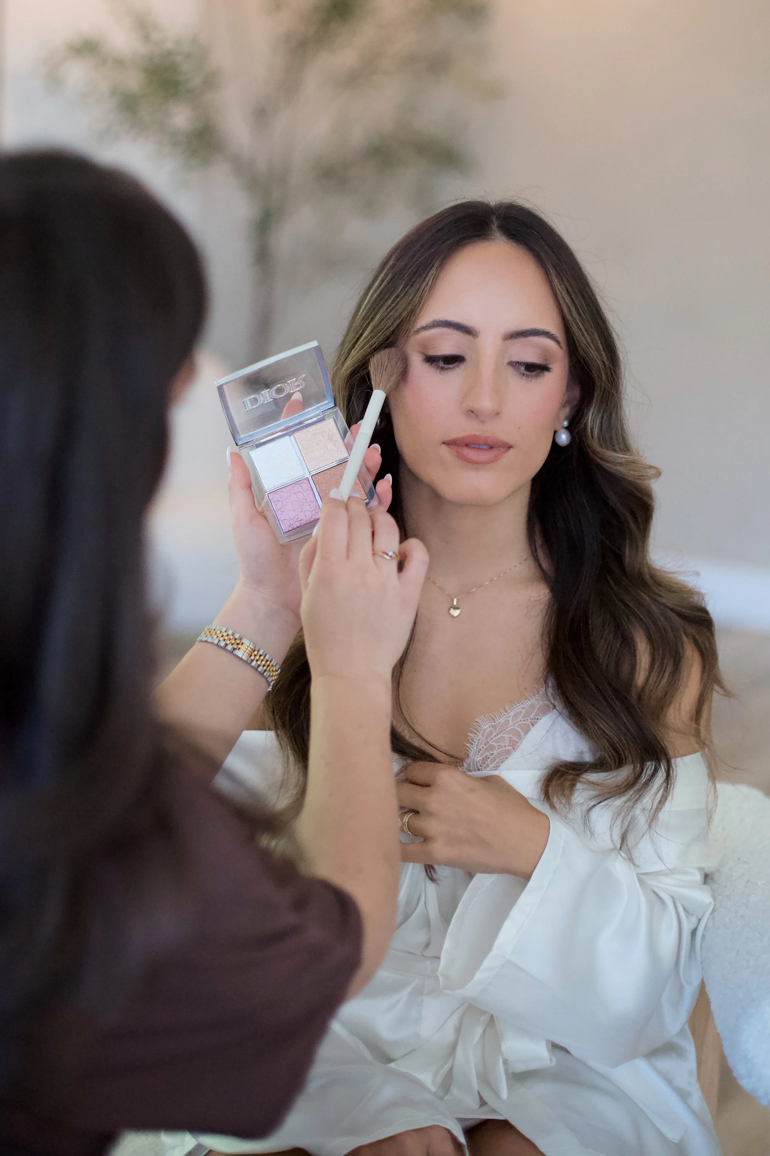 A woman with long brown hair getting her makeup done by another woman who is applying eyeshadow with a brush. The woman receiving makeup is wearing a white off-shoulder top and jewelry, and the makeup artist is holding a palette of four eyeshadow sha