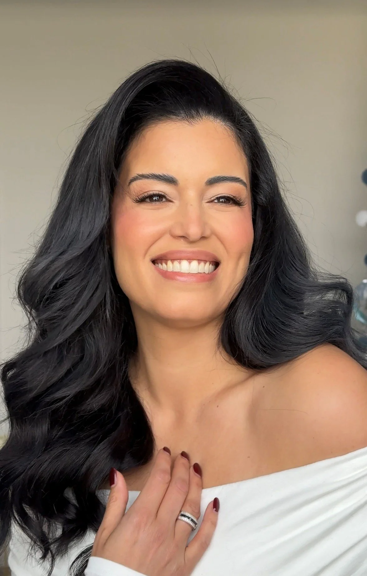 A woman with long black hair, smiling, wearing an off-shoulder white top, showing her diamond ring and painted nails.
