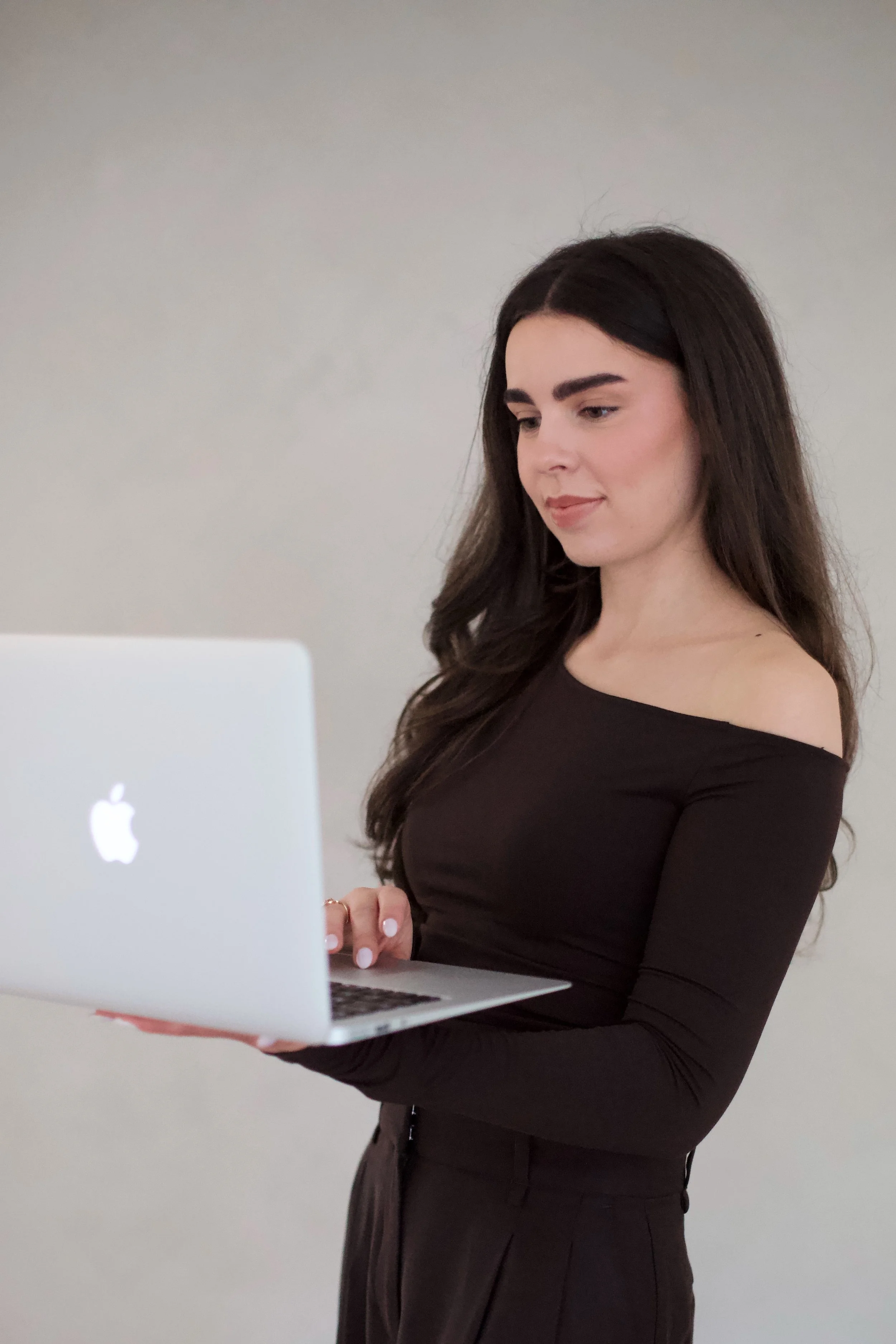 A young woman with long dark hair, wearing a black off-shoulder top and black pants, holding an Apple MacBook, standing against a plain light-colored wall.
