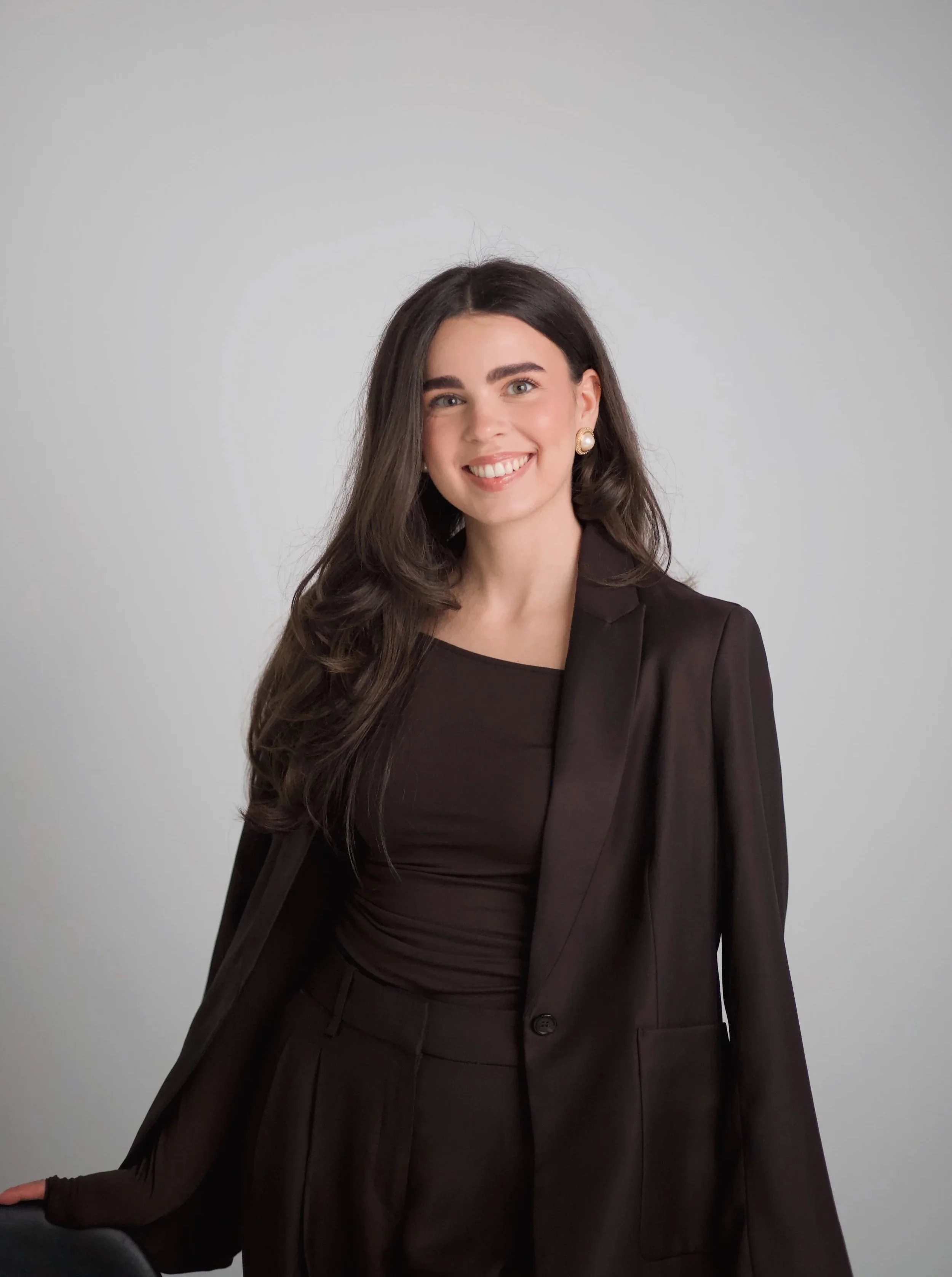 A smiling woman with long dark hair wearing a black suit and pearl earrings, standing against a light gray background.