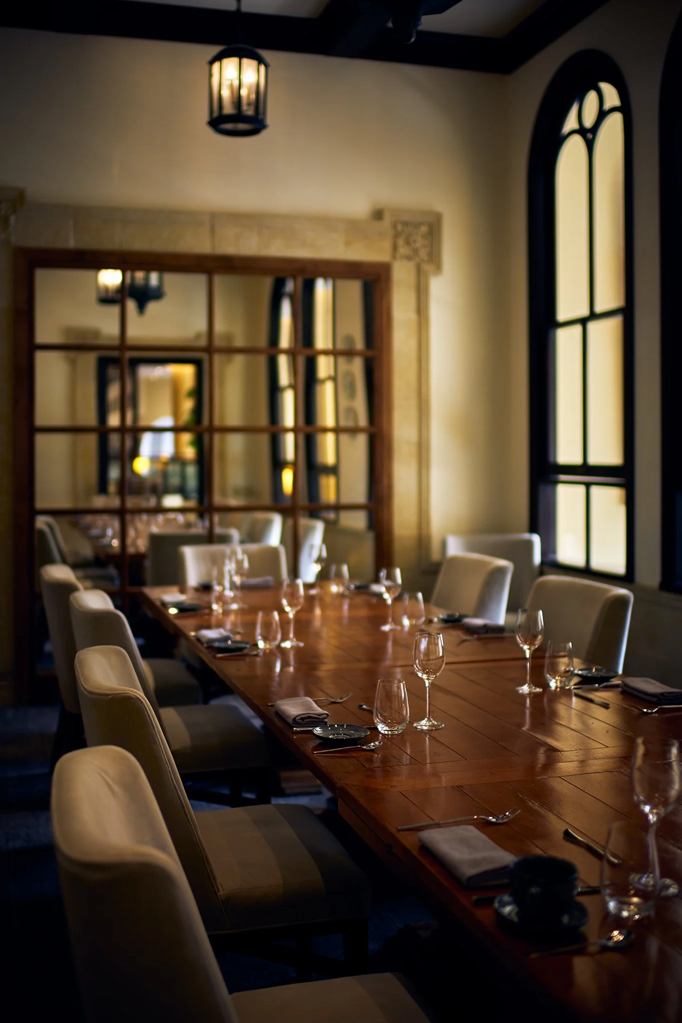 A dining room with a long wooden table set with wine glasses, silverware, napkins, and black dishes, surrounded by beige upholstered chairs. The room has large arched windows and a wooden-framed mirror reflecting the interior, with a lantern-style ceiling light.