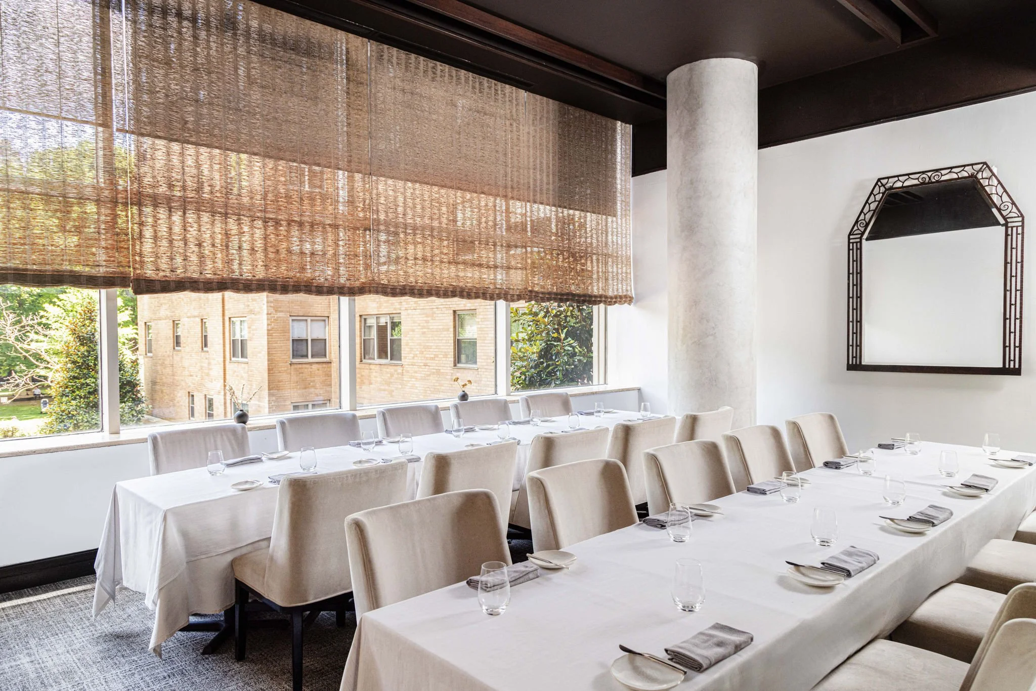 An elegant dining room with two long tables covered in white tablecloths, set with glasses, plates, and napkins. Large windows with woven shades allow natural light, revealing a view of brick buildings and greenery outside. There is a white column, a decorative mirror on the wall, and dark ceiling beams.