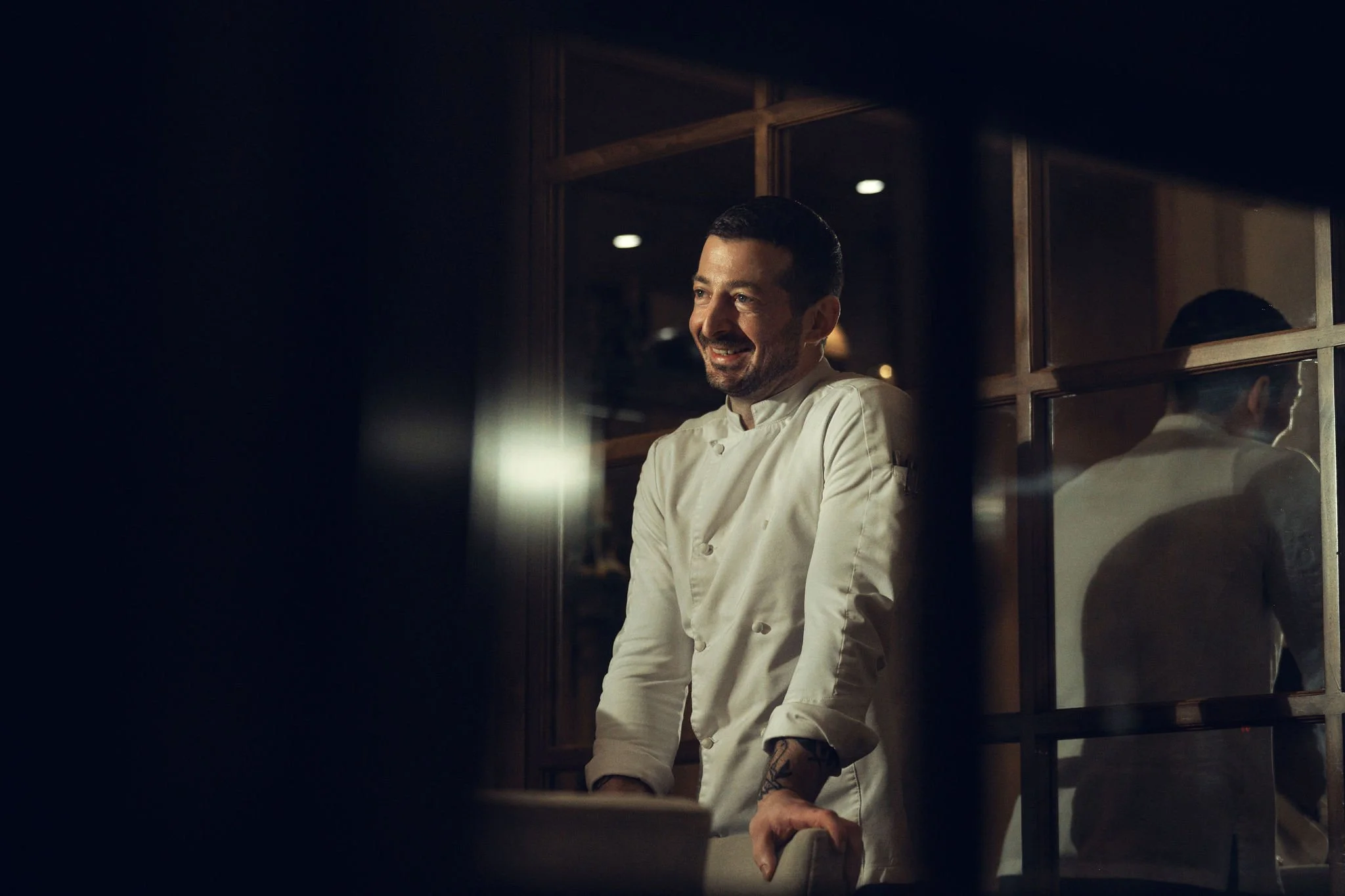 Smiling chef standing near a wooden window reflection in a dimly lit kitchen.