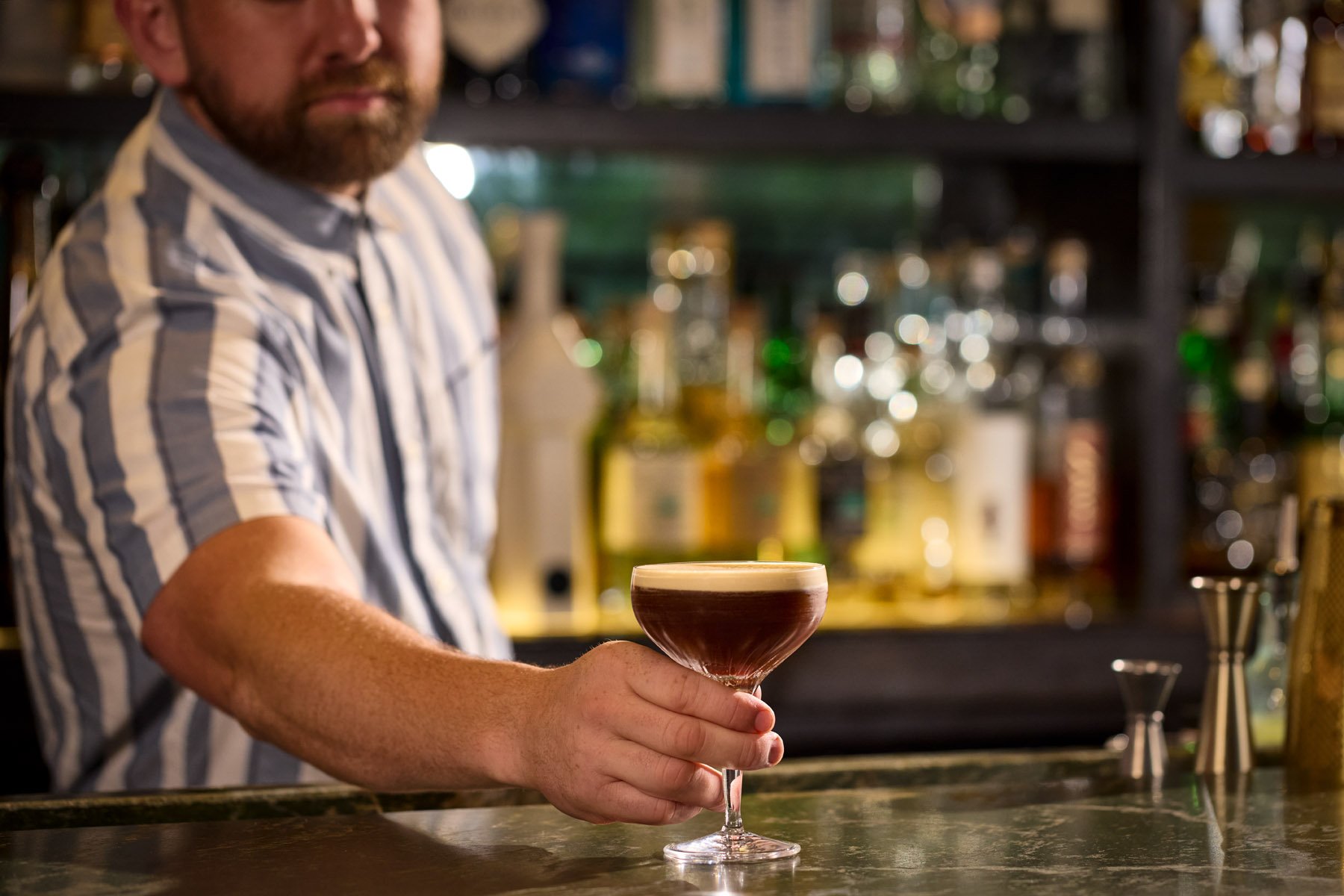 A bartender holding a cocktail in a coupe glass at a bar with bottles of alcohol on shelves behind him.